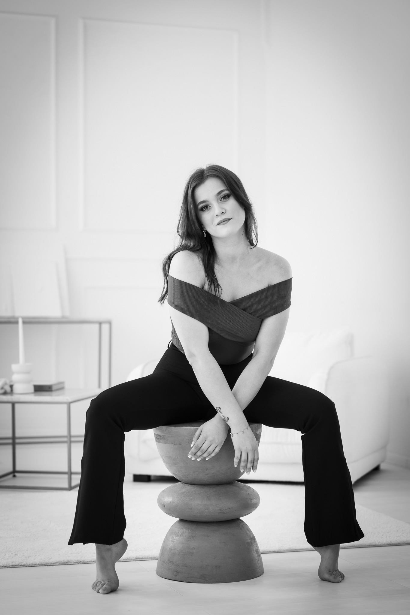 Czarno-białe zdjęcie kobiety siedzącej na drewnianym stołku w kształcie kamieni, ubrana w bluzkę z odkrytymi ramionami i szerokie spodnie, z tatuażem na przedramieniu i kolczykami kołami,...