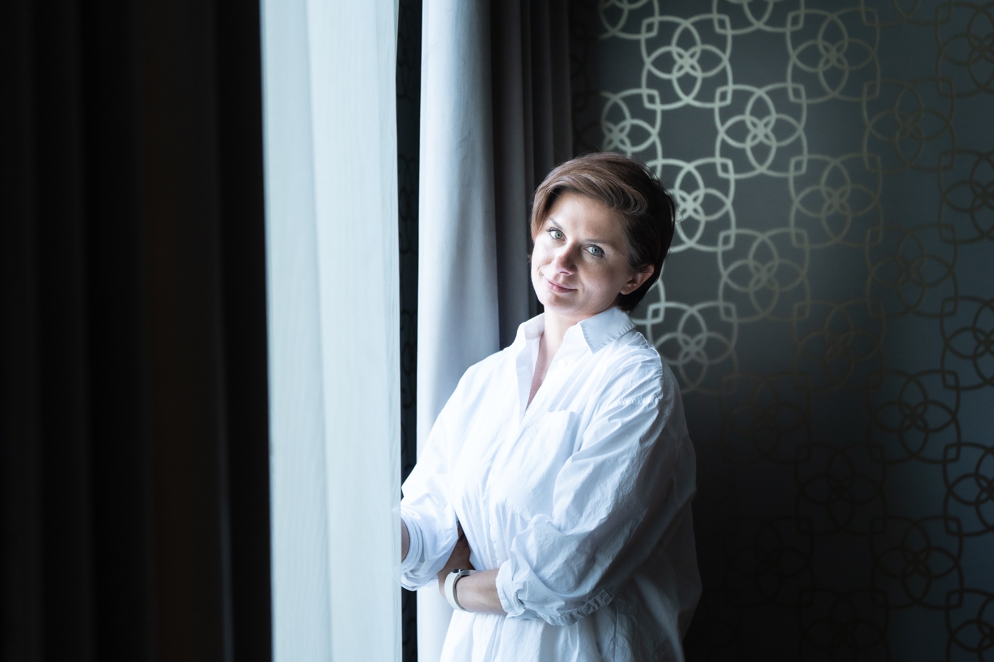 Kobieta w białej koszuli stojąca przy oknie z firankami, na tle ściany z geometrycznym wzorem, delikatny uśmiech.