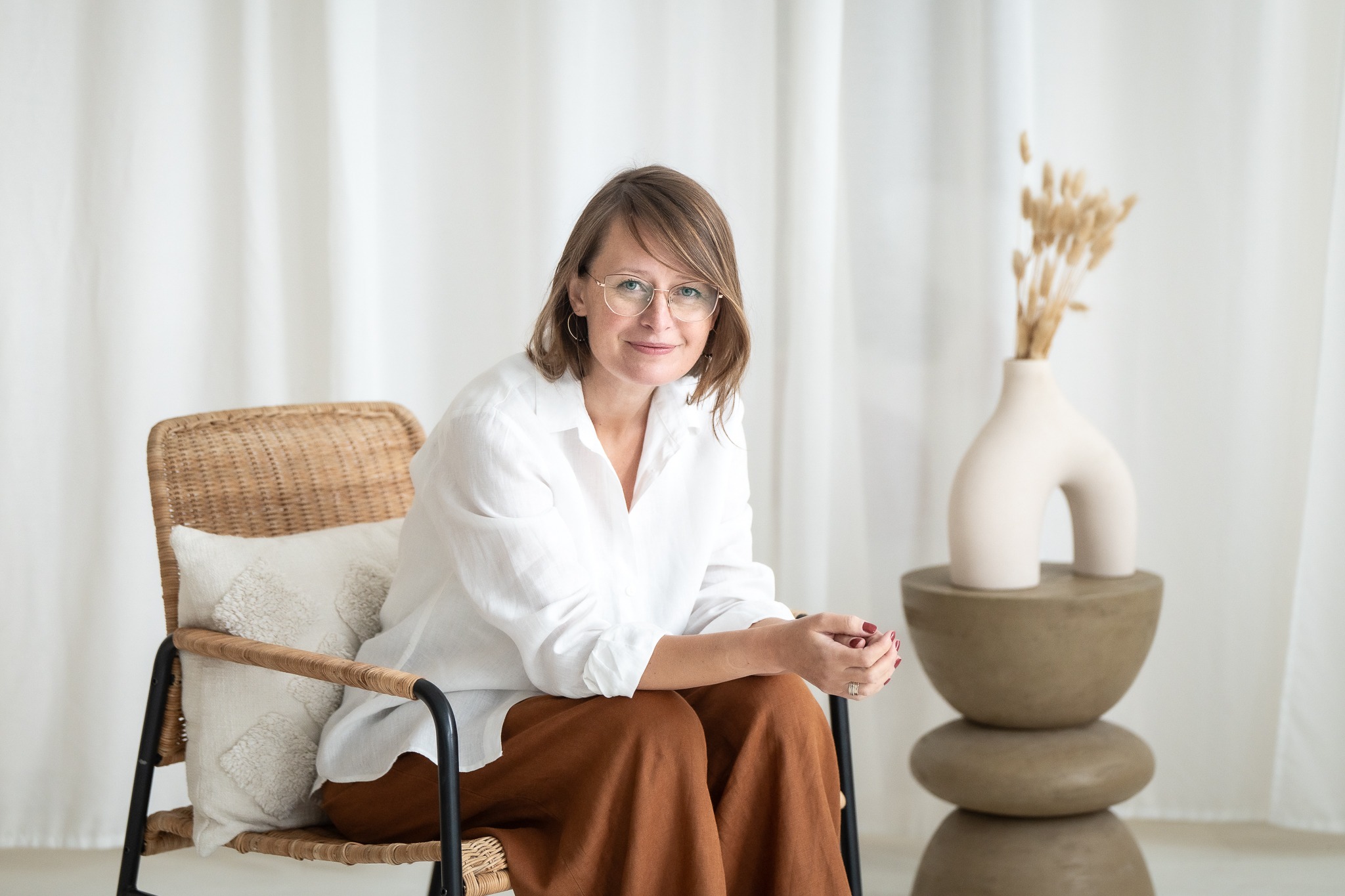 Portret kobiety w okularach, siedzącej na wiklinowym krześle z poduszką, w białej koszuli i brązowych spodniach, z dekoracyjnym wazonem z suszem w tle, w jasnym wnętrzu.