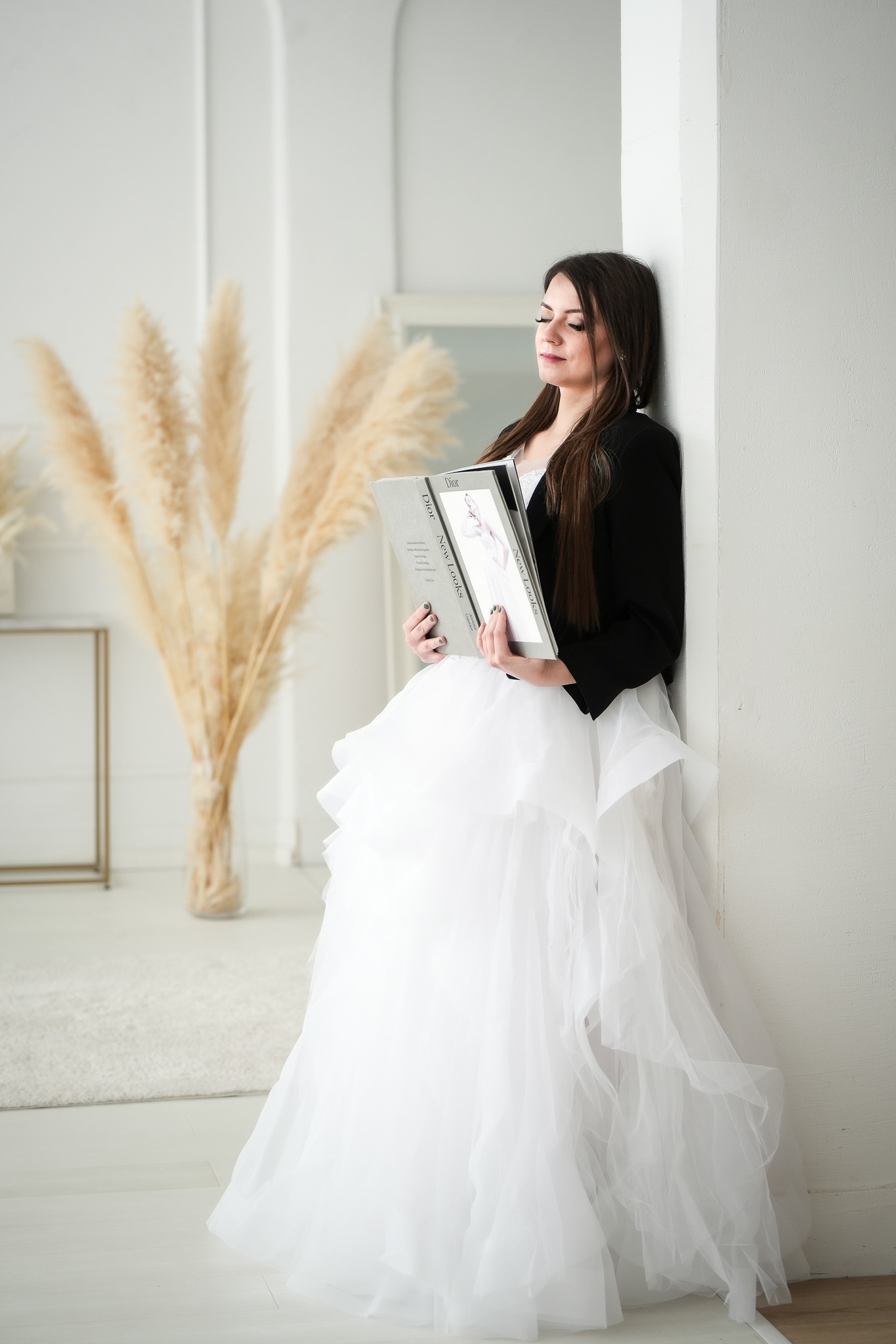 Kobieta w białej, warstwowej sukni i czarnej marynarce opiera się o ścianę, trzymając w rękach książkę 'Dior New Looks' w jasnym pomieszczeniu z dekoracyjną trawą pampasową.