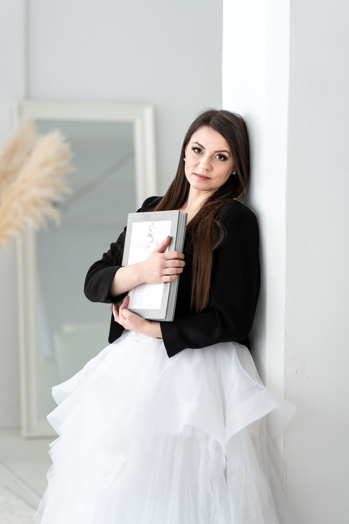 Kobieta w białej sukni i czarnej marynarce opiera się o białą ścianę, trzymając w rękach książkę Dior. W tle widoczne lustro i dekoracyjne trawy.