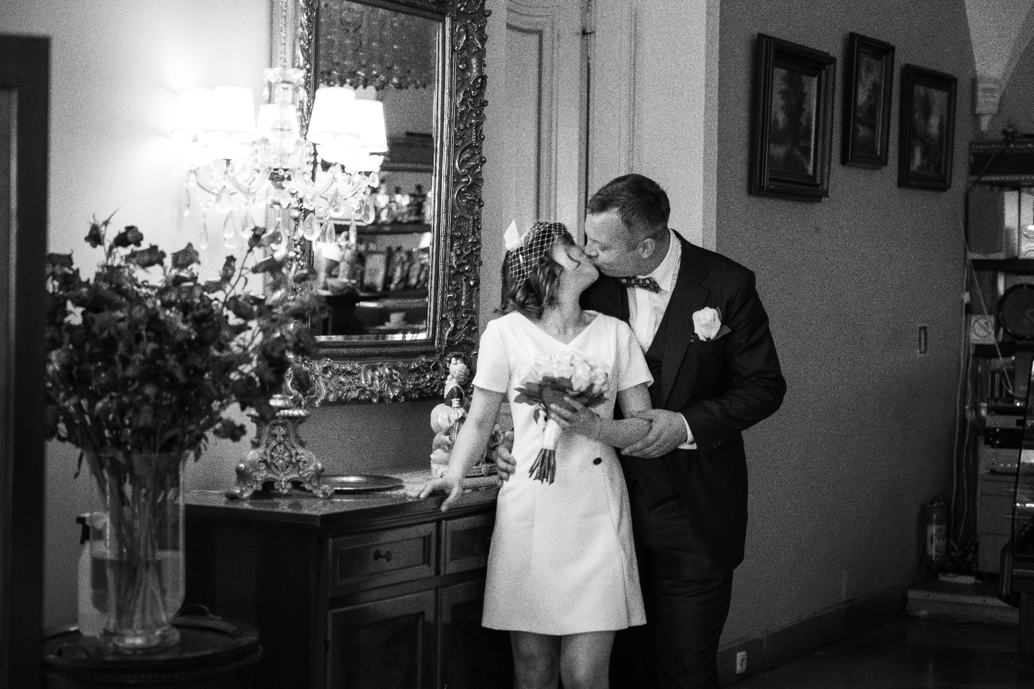 Czarno-białe zdjęcie pary młodej podczas pocałunku w eleganckim wnętrzu, z ozdobnym lustrem, żyrandolem i bukietem kwiatów.