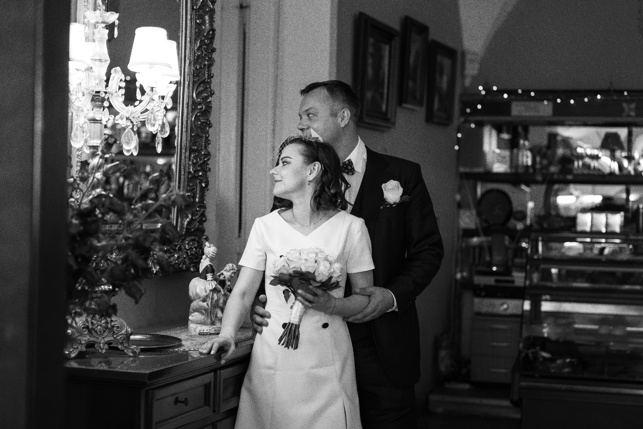Czarno-białe zdjęcie pary młodej: panna młoda w prostej sukience z bukietem białych róż, pan młody w ciemnym garniturze z muszką i różą w butonierce, stoją obok siebie w eleganckim wnętrzu...