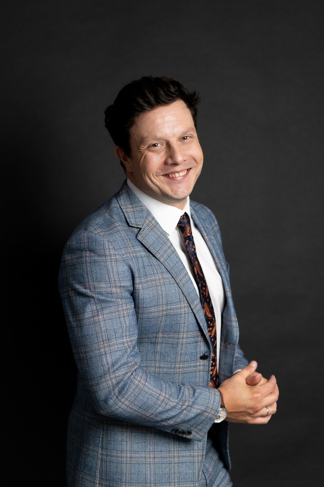 Portret uśmiechniętego mężczyzny w błękitnej marynarce w kratę i wzorzystym krawacie na ciemnym tle, z widocznym zegarkiem na nadgarstku.