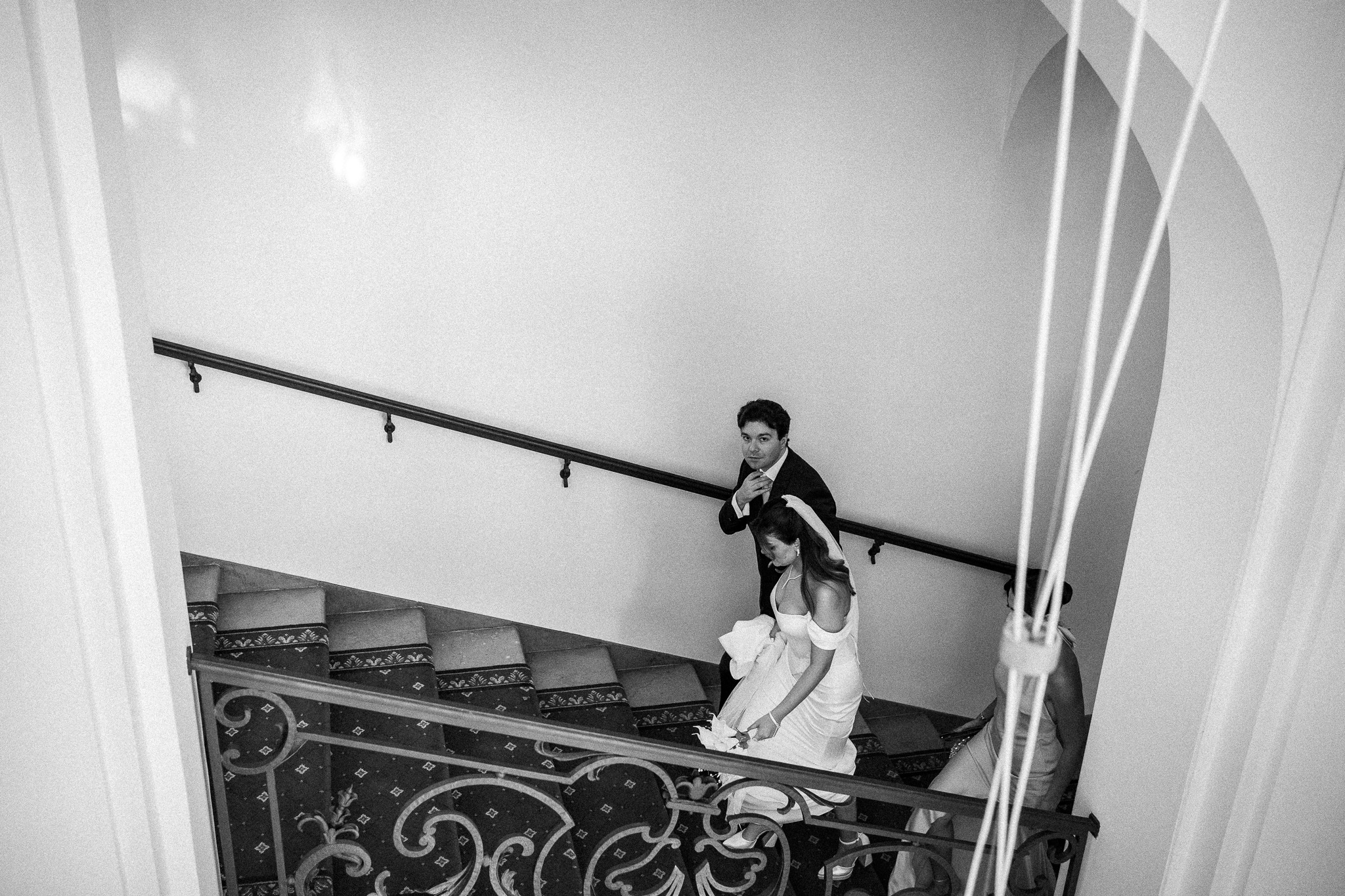 Czarno-białe zdjęcie pary młodej schodzącej po schodach z ozdobną balustradą. Pan młody w garniturze, panna młoda w sukni ślubnej. Ujęcie z góry, widoczne detale schodów.