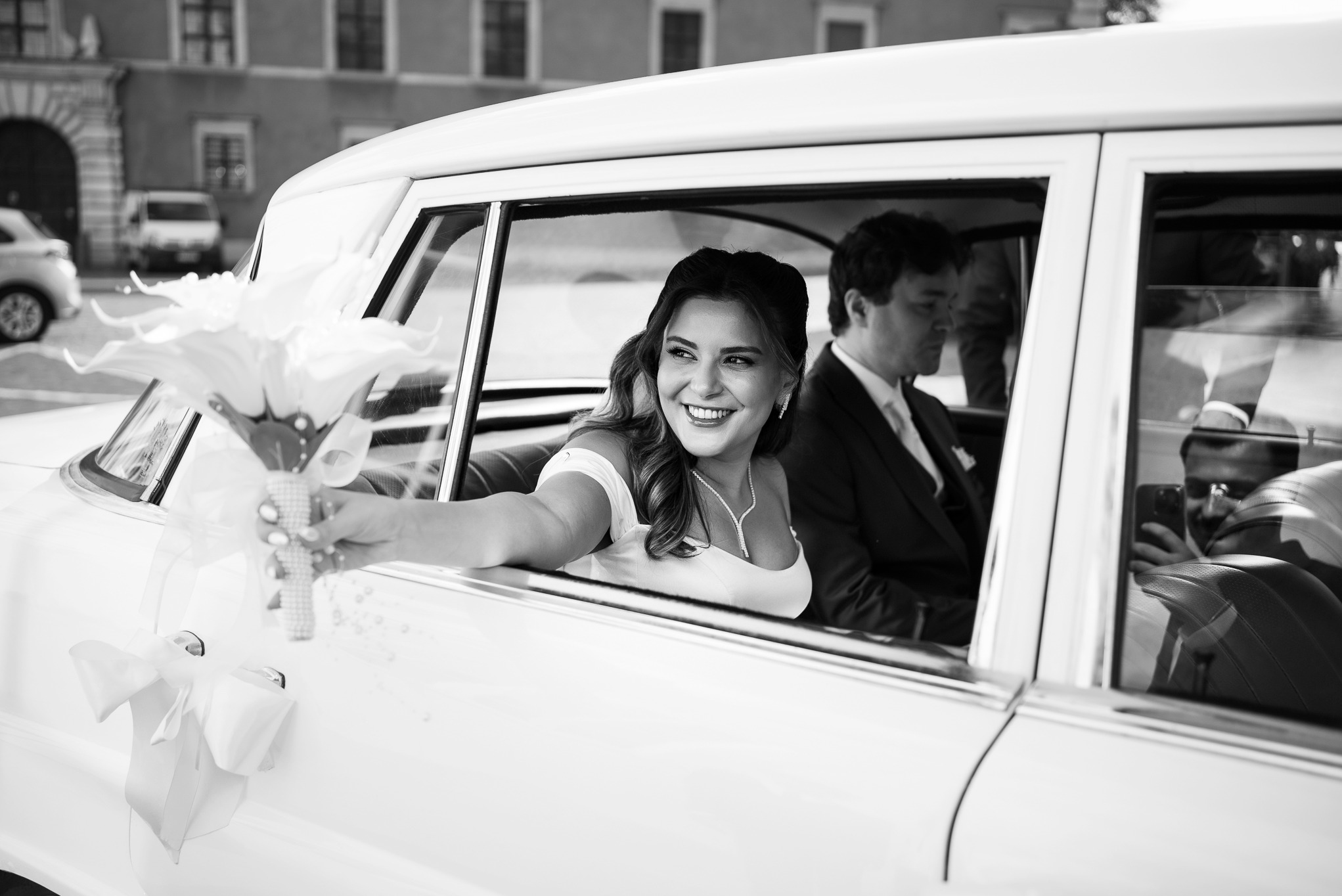Uśmiechnięta panna młoda w białej sukni, wyciąga bukiet z okna białej limuzyny. Obok siedzi pan młody w garniturze. Czarno-biała fotografia ślubna.