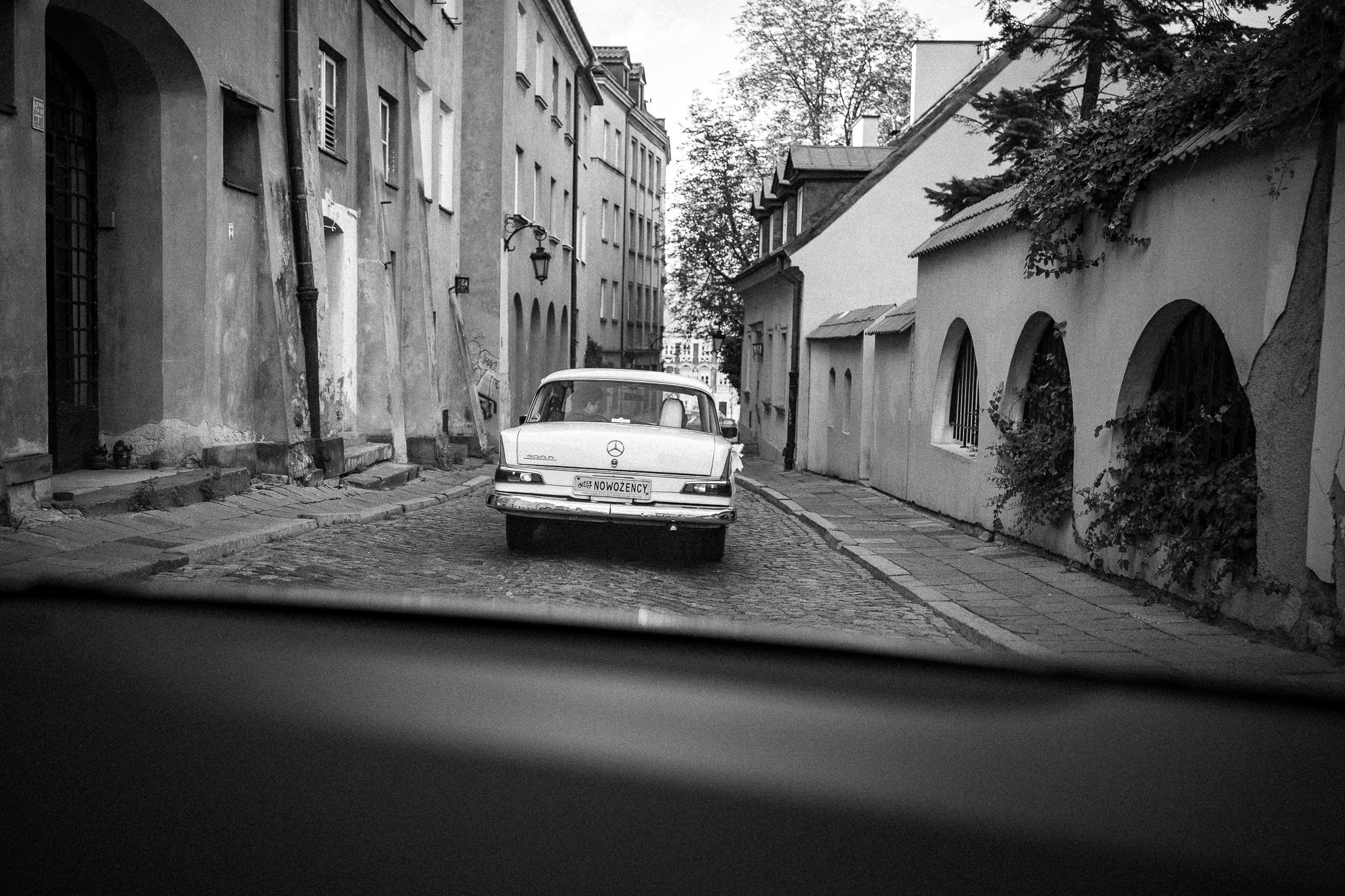 Czarno-białe zdjęcie zza szyby samochodu: Mercedes 200D na brukowanej uliczce Starego Miasta, w tle kamienice i drzewa. Tablica rejestracyjna z napisem 'NOWOŻENCY'.