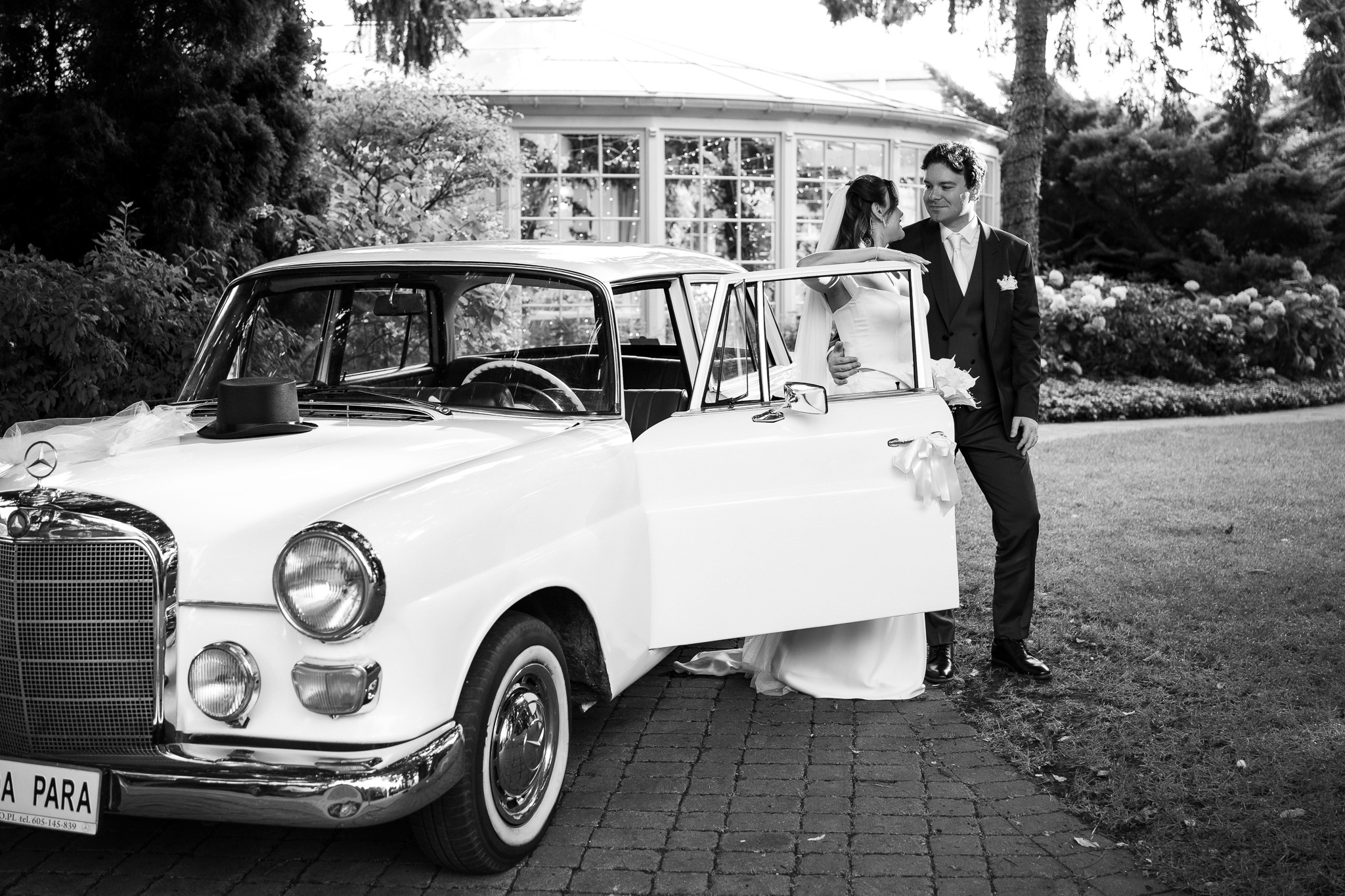 Czarno-białe zdjęcie pary młodej przy białym, zabytkowym Mercedesie z otwartymi drzwiami, na tle ogrodu i oranżerii. Na masce auta cylinder i woalka.