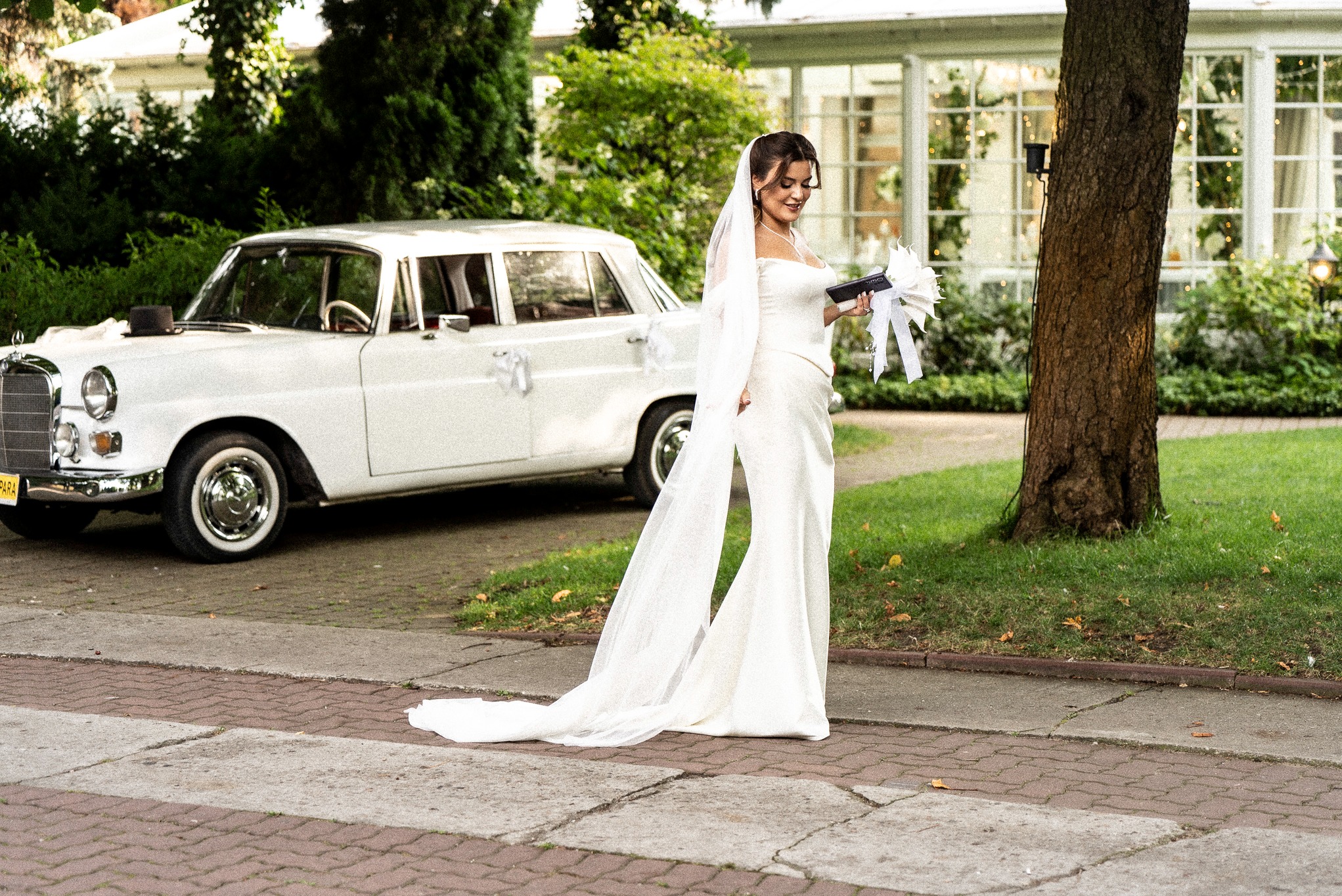 Panna młoda w białej sukni i welonie, trzymająca bukiet, pozuje obok białego, zabytkowego Mercedesa z dekoracjami ślubnymi na tle zieleni i budynku.