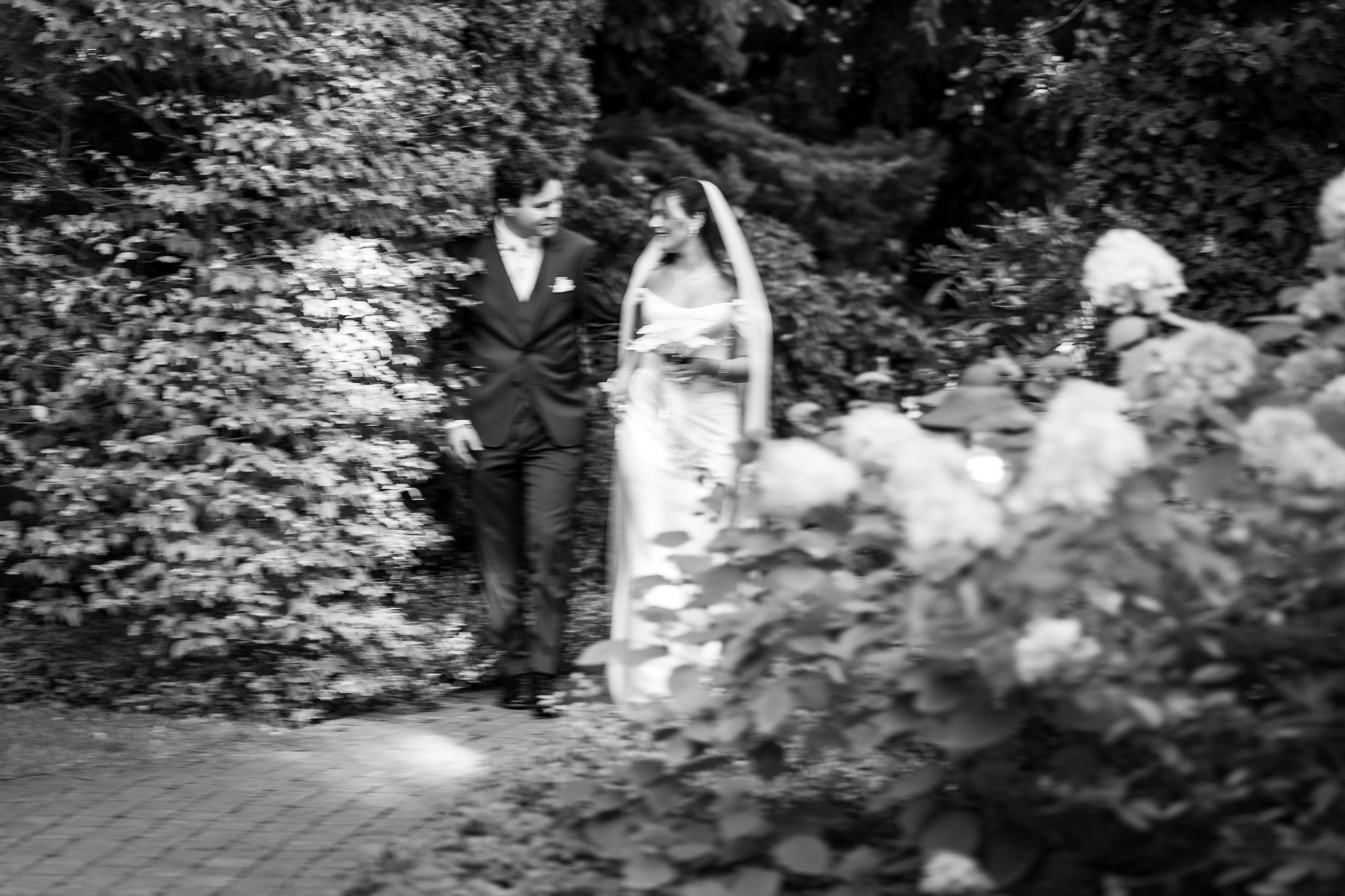 Czarno-białe zdjęcie pary młodej spacerującej w Łodzi, otoczonej zielenią. Panna młoda w sukni i welonie, pan młody w garniturze. Ujęcie w ruchu, z rozmytym pierwszym planem.