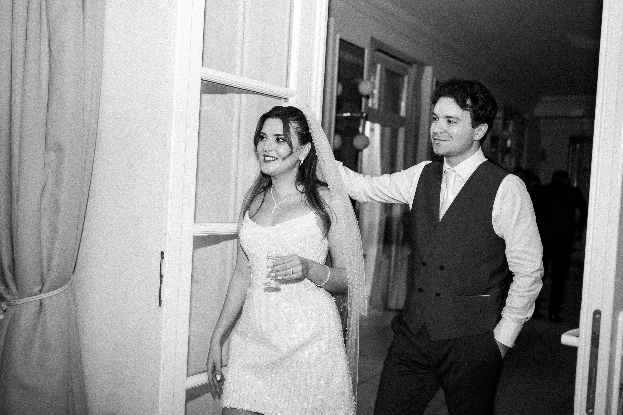 Czarno-białe zdjęcie pary młodej: panna młoda w krótkiej sukience i welonie, trzyma kieliszek, obok pan młody w kamizelce, oboje uśmiechnięci, w tle drzwi.