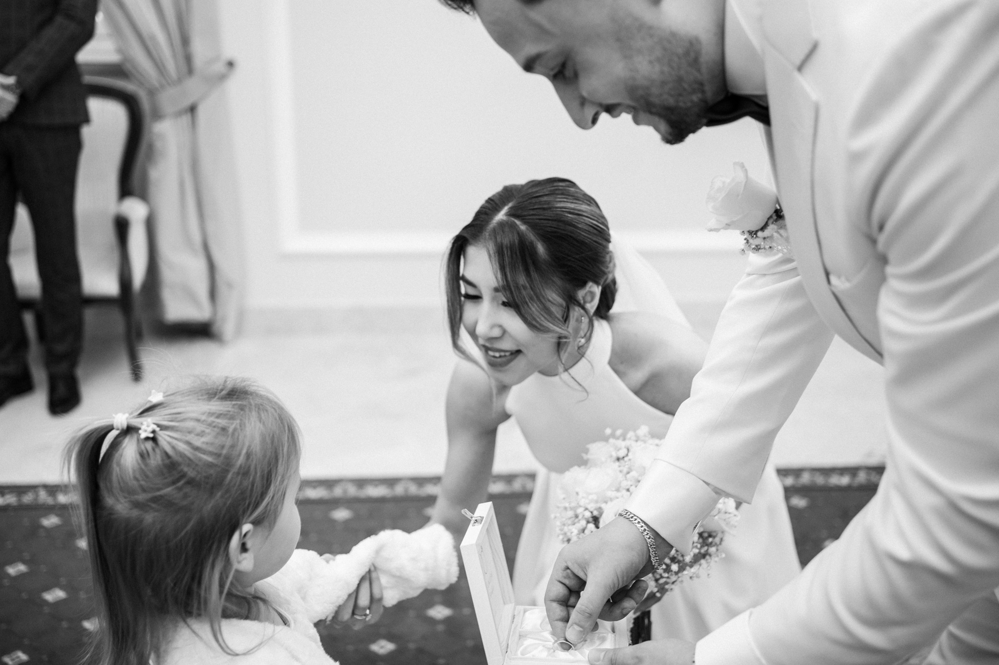 Czarno-białe zdjęcie z ceremonii ślubnej: panna młoda uśmiecha się, gdy pan młody wyjmuje obrączkę z pudełka, a mała dziewczynka obserwuje z bliska.