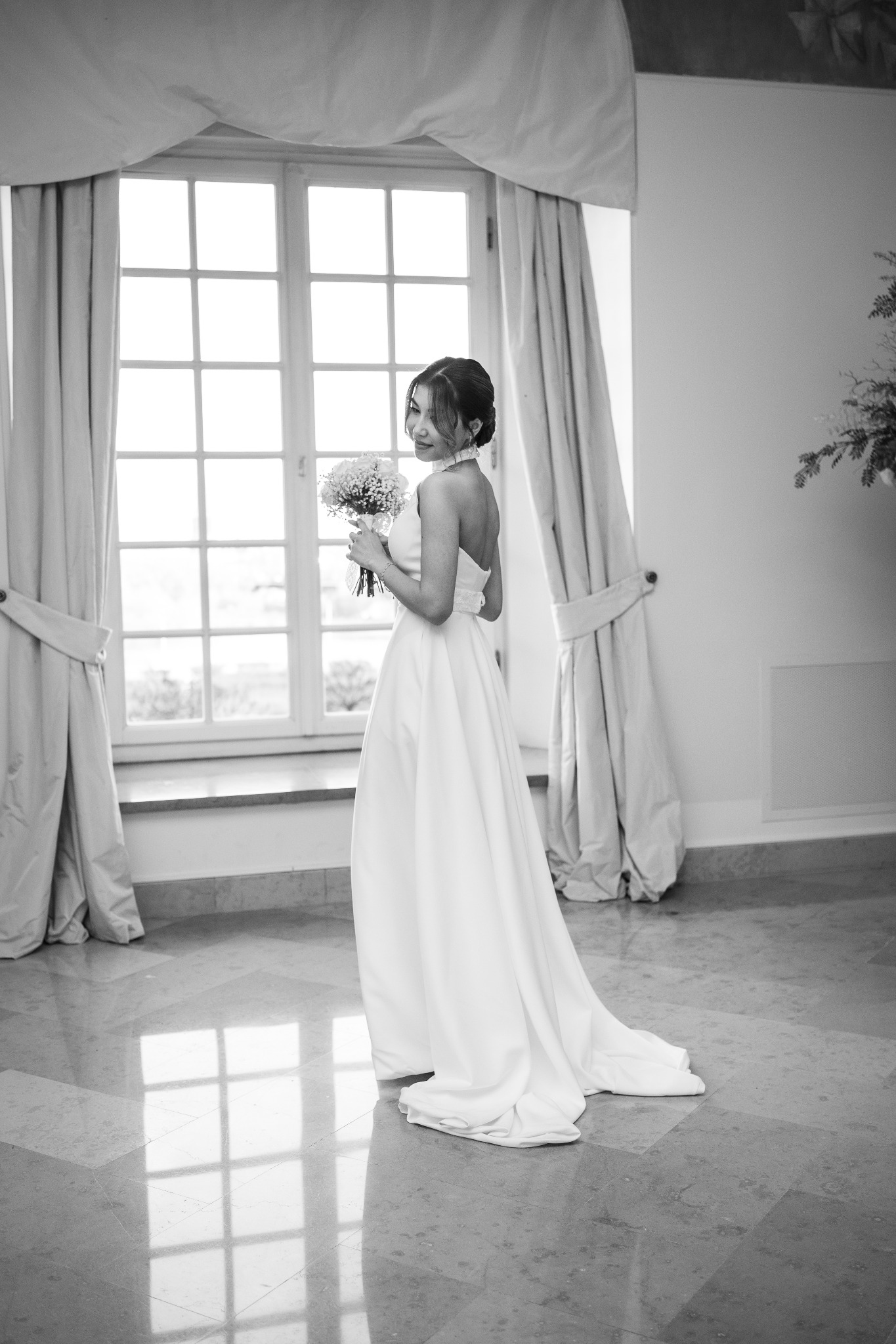Elegancka panna młoda w białej sukni, z bukietem, spogląda przez ramię w jasnym wnętrzu z dużym oknem. Zdjęcie w czerni i bieli, podkreślające subtelność i klasę.