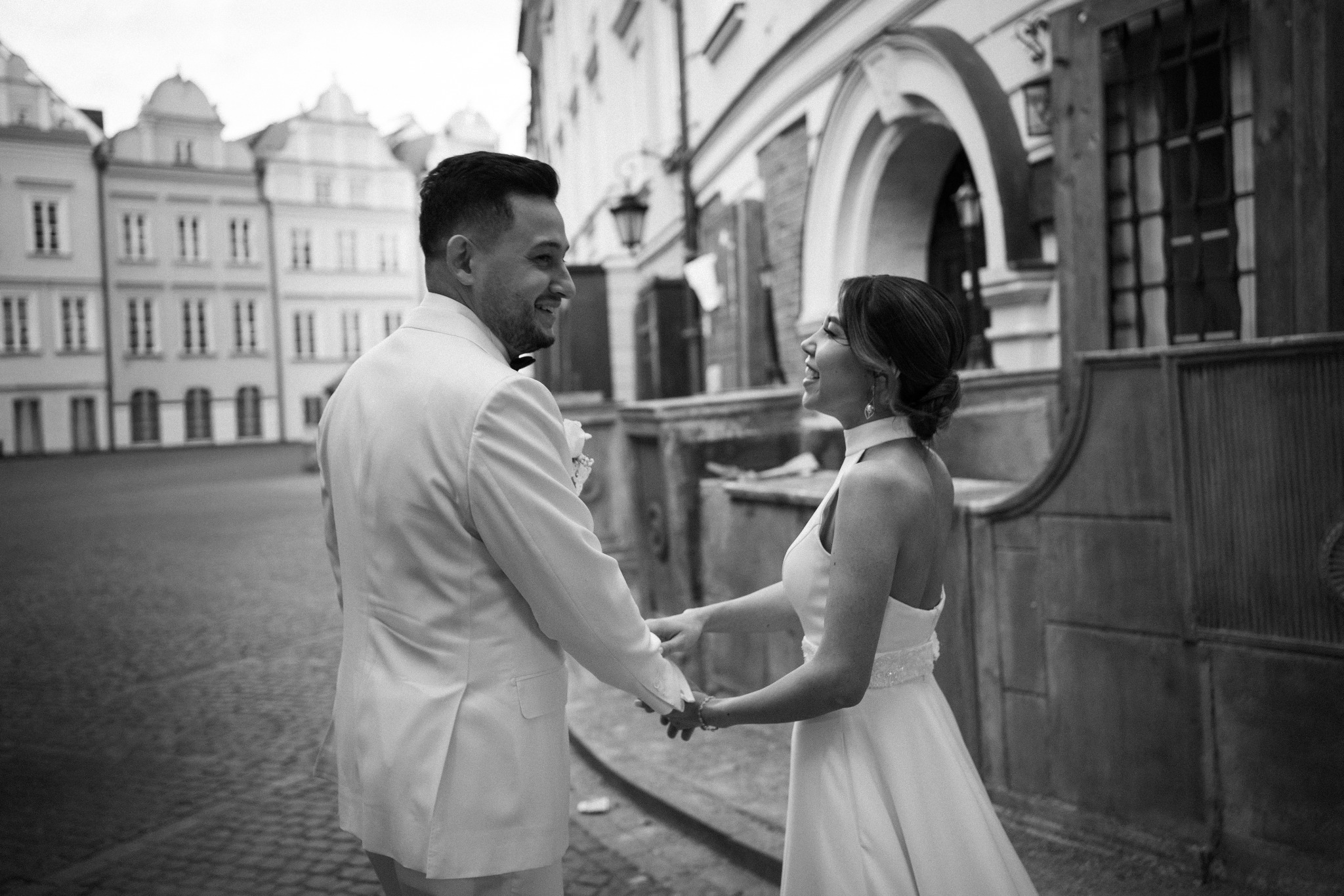 Czarno-białe zdjęcie uśmiechniętej pary młodej trzymającej się za ręce na tle architektury miejskiej z arkadami i budynkami w tle.