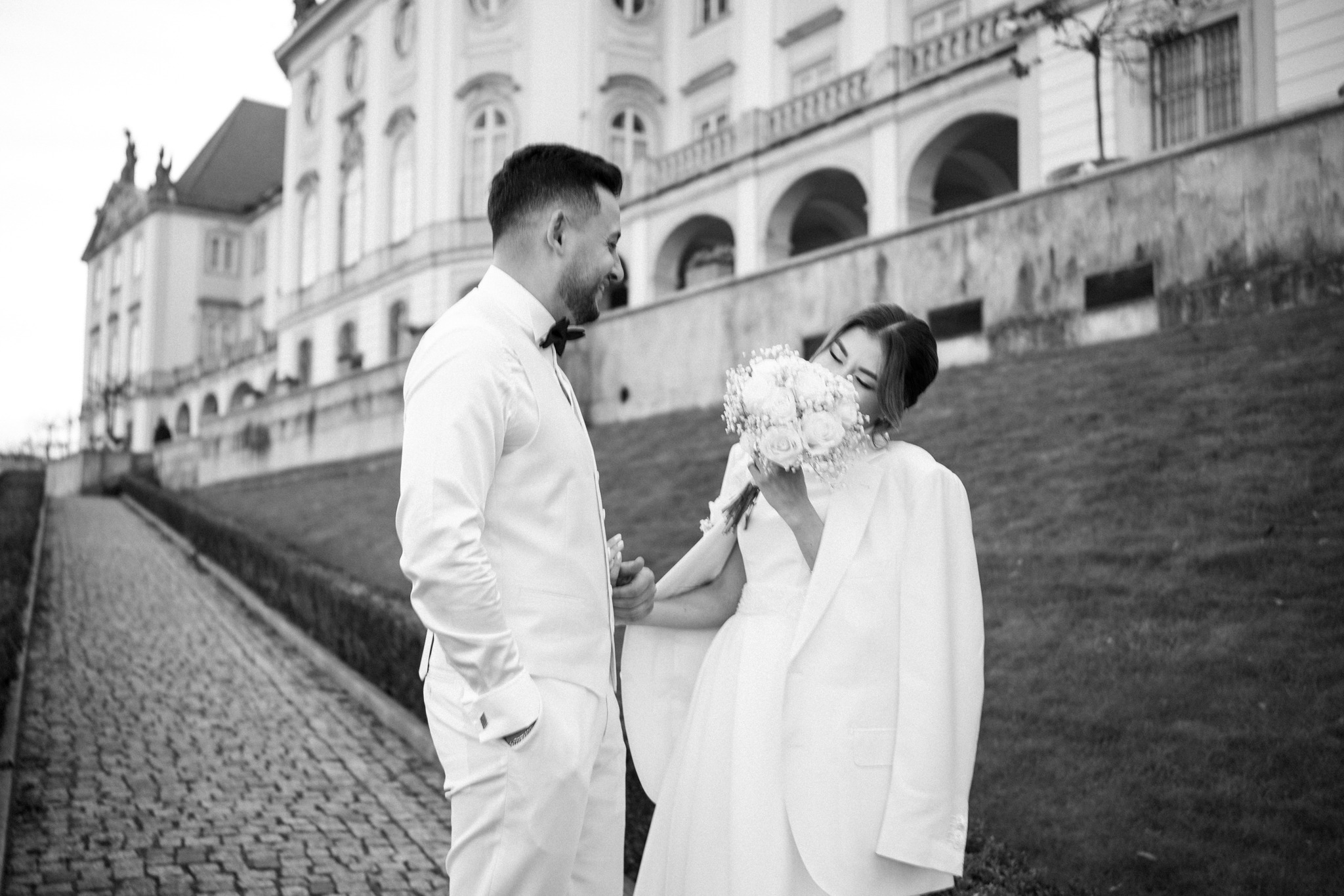 Czarno-białe zdjęcie uśmiechniętej pary młodej w białych strojach, panna młoda zakrywa twarz bukietem róż, w tle pałac i brukowana droga.