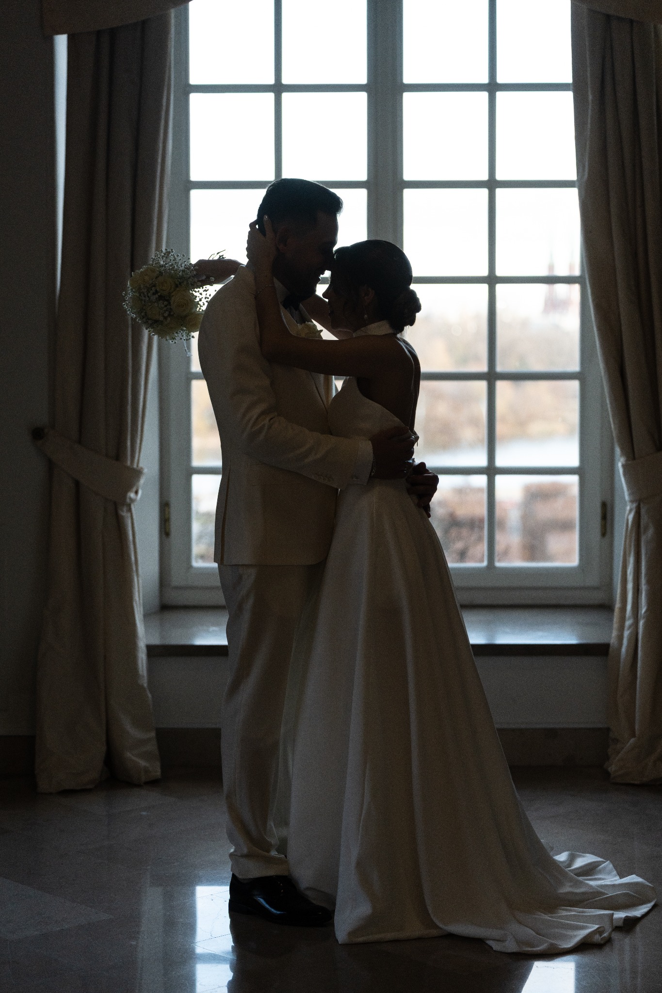 Silueta pary młodej w objęciach na tle okna z widokiem na zewnątrz. Ona w sukni ślubnej, on w jasnym garniturze. Delikatny bukiet w dłoni. Romantyczna atmosfera.