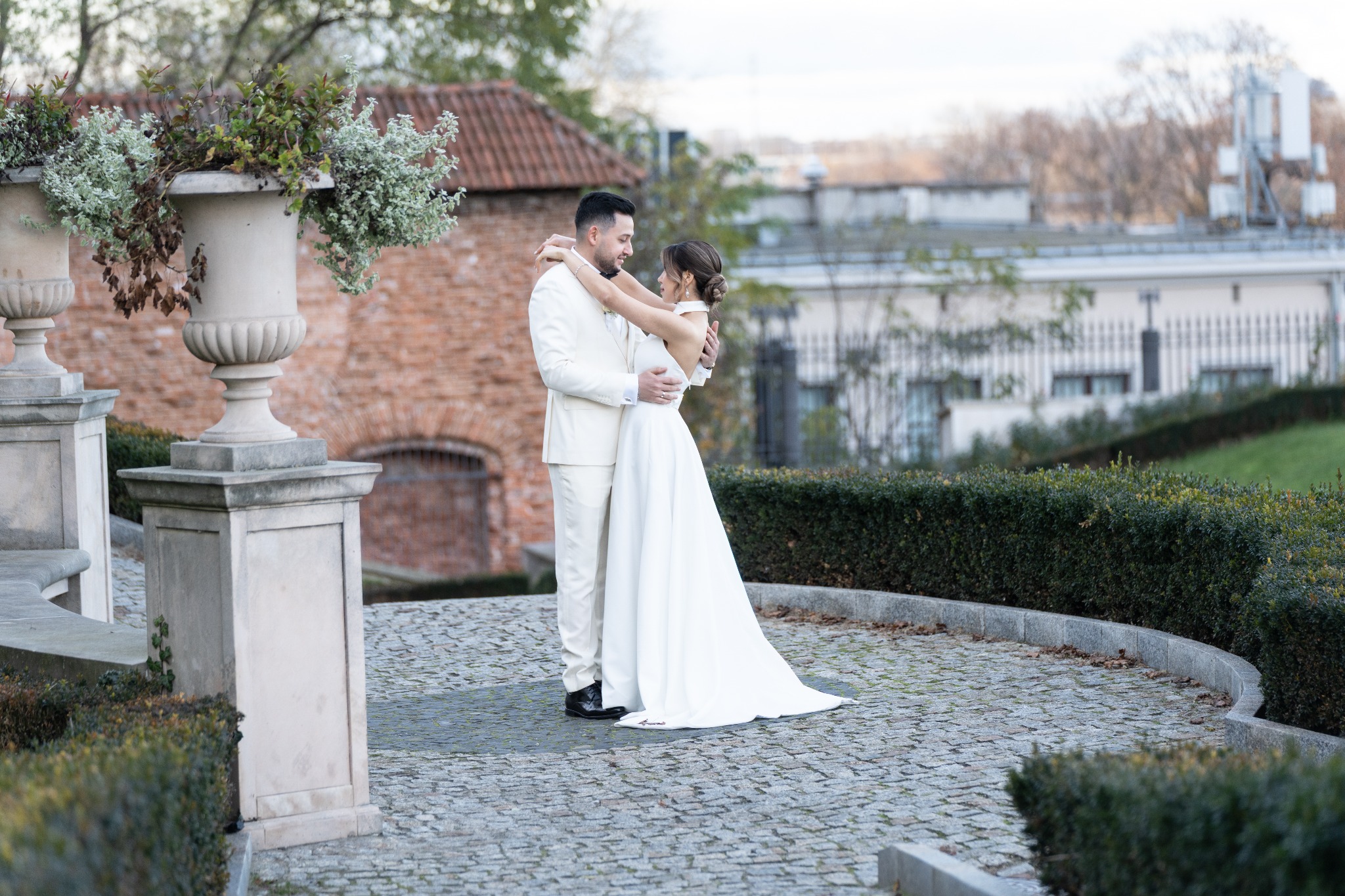 Para młoda w objęciach na tle zabytkowej architektury i zieleni. Panna młoda w białej sukni, pan młody w kremowym garniturze. Ujęcie z delikatnym rozmytym tłem.