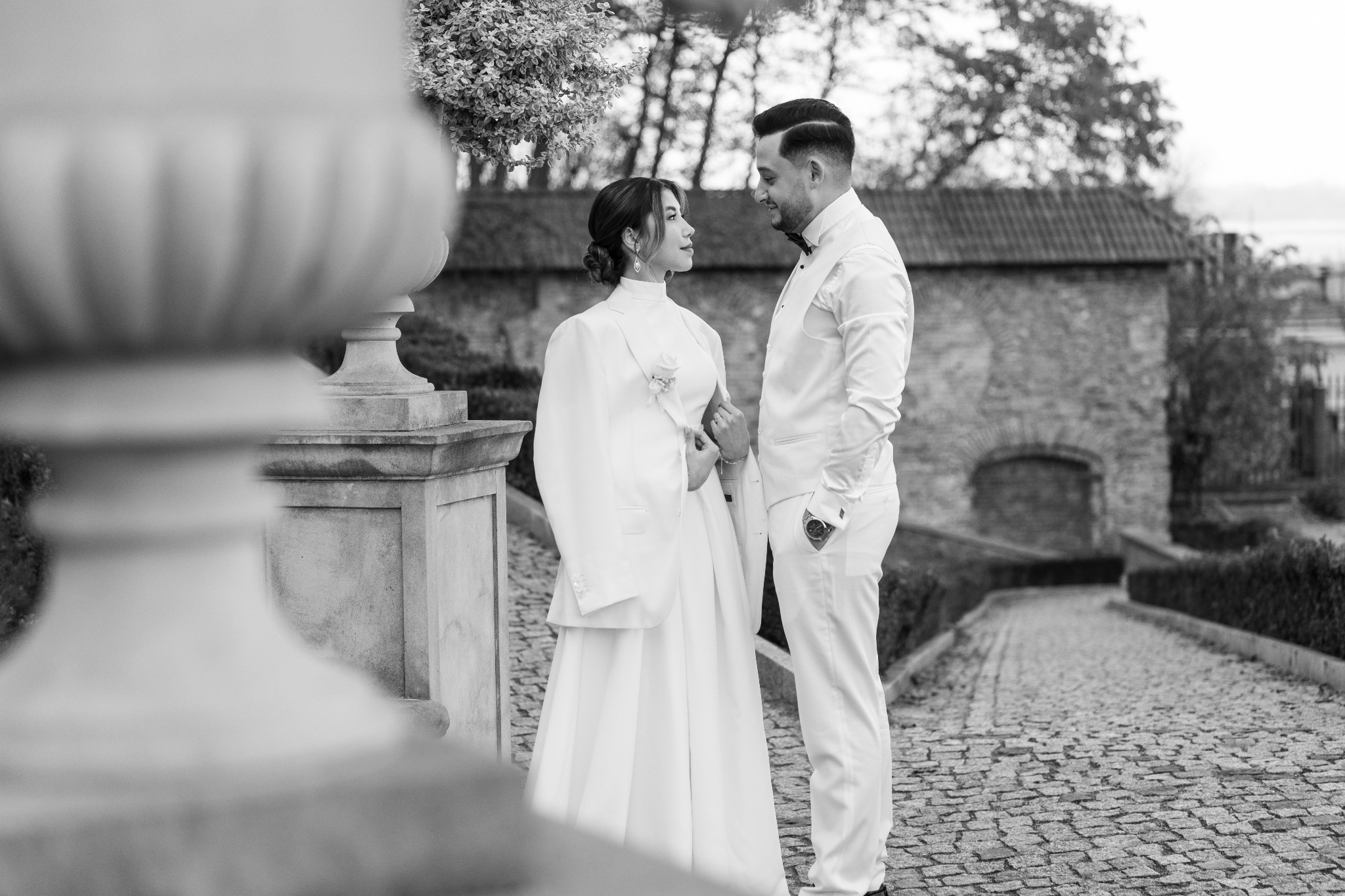 Czarno-białe zdjęcie pary młodej w eleganckich, białych strojach na tle kamiennej ścieżki i zabytkowej architektury. Ona w sukni i marynarce, on w smokingu.