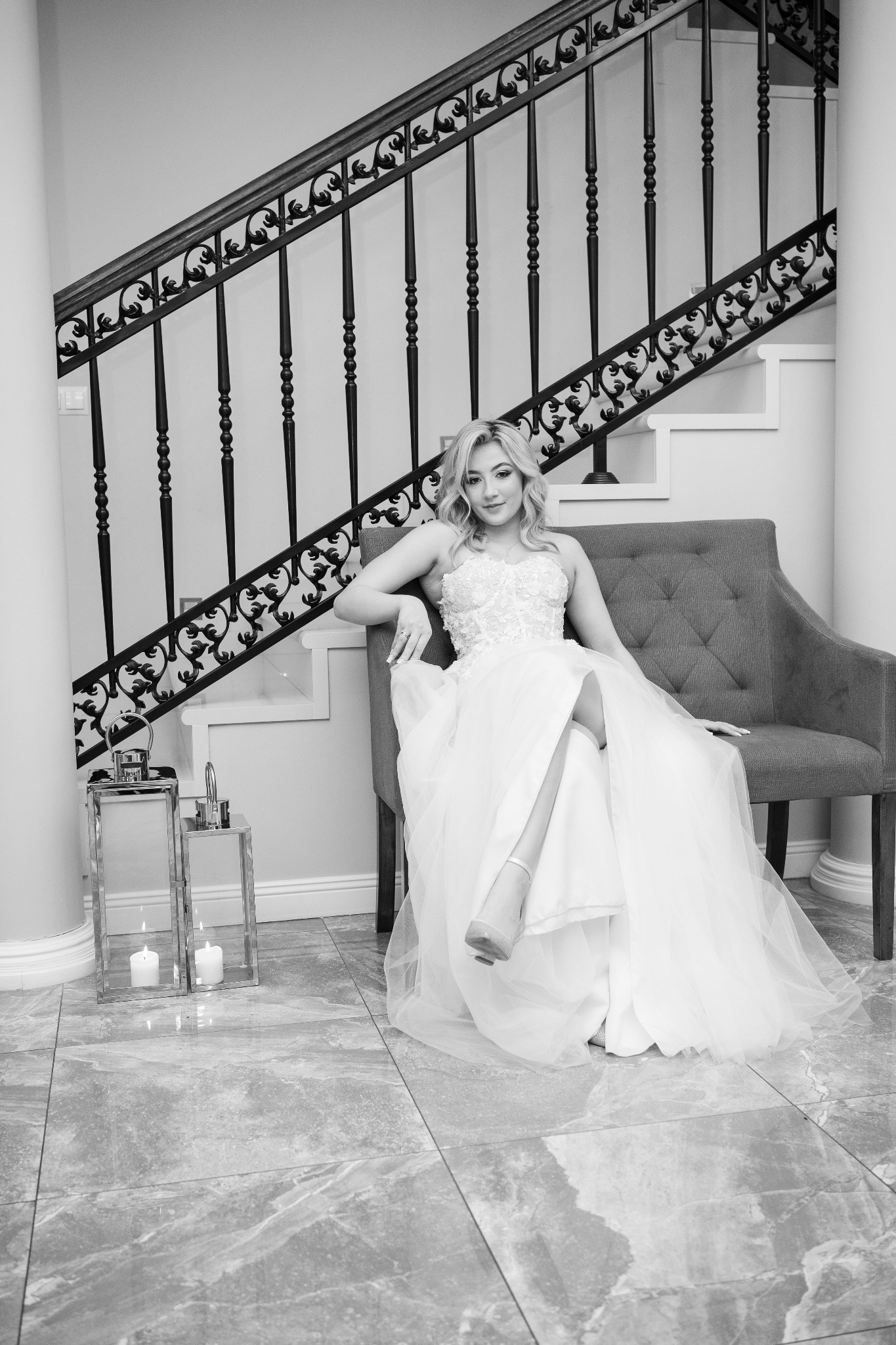 Elegancka sesja w czerni i bieli: Panna młoda w koronkowej sukni pozuje siedząc na sofie na tle schodów z ozdobną balustradą. Obok lampiony ze świecami.
