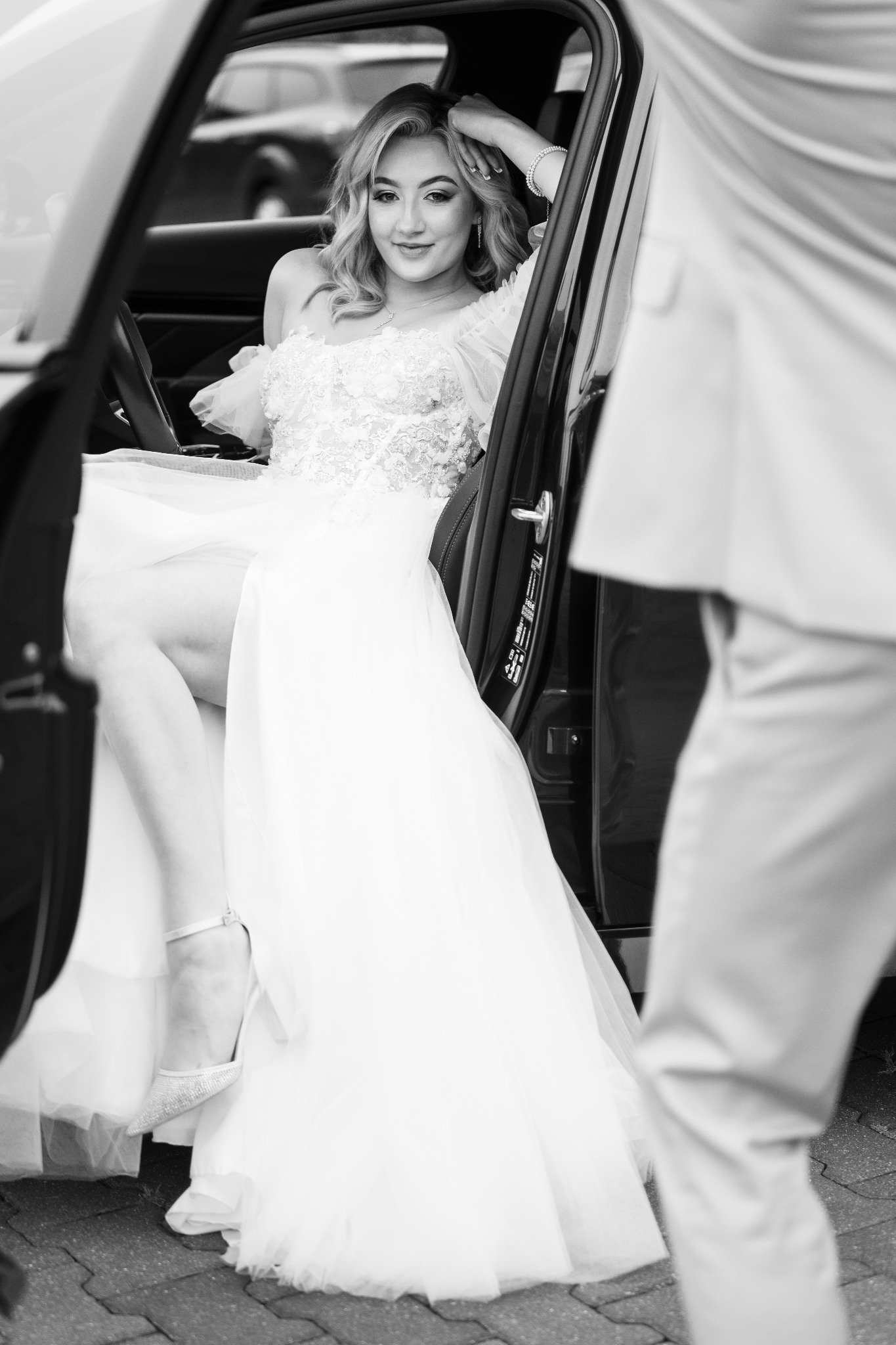 Czarno-białe zdjęcie panny młodej w sukni ślubnej, siedzącej w samochodzie z otwartymi drzwiami, z widoczną nogą i butem. Obok stoi mężczyzna w garniturze.