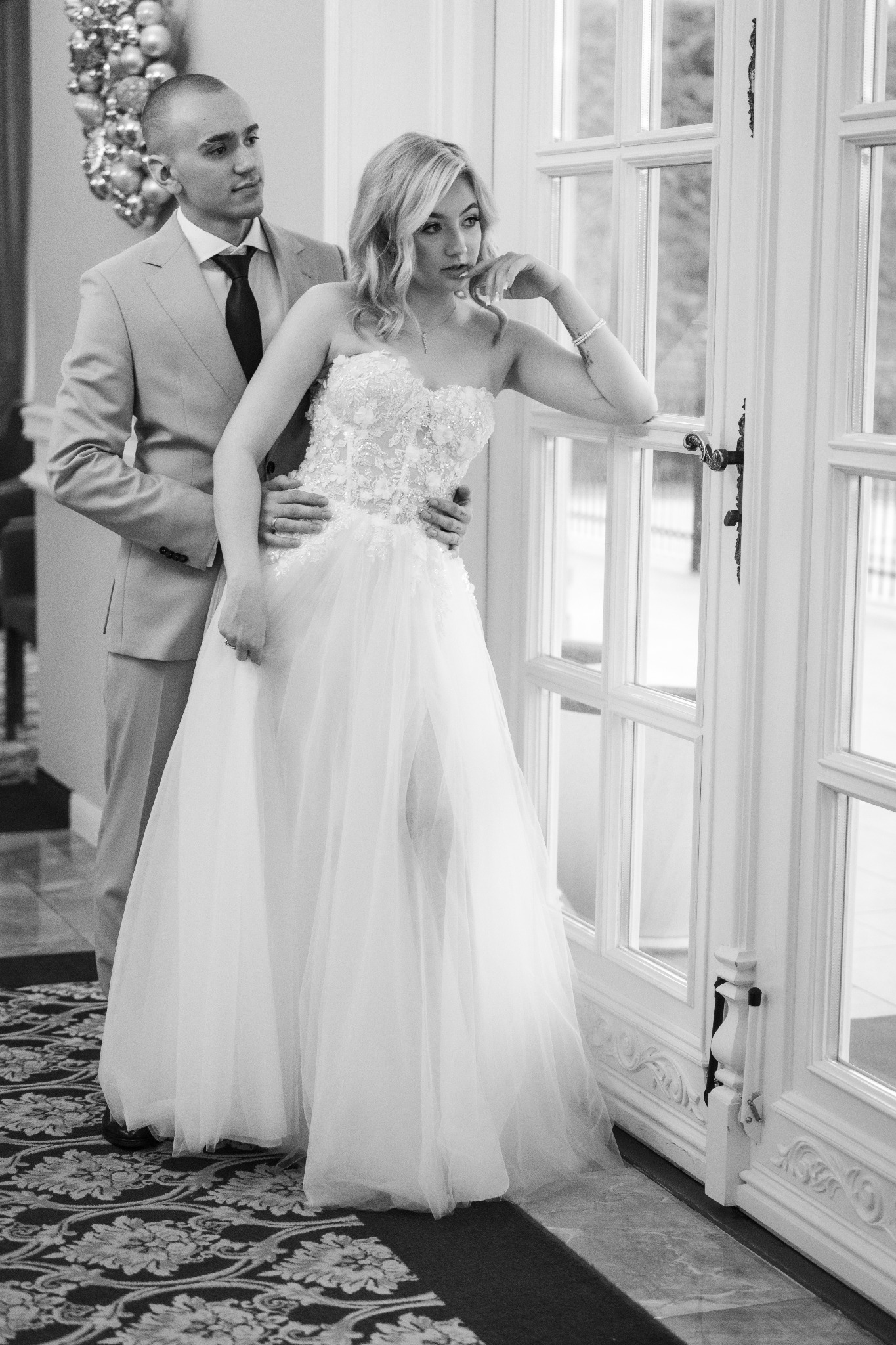 Czarno-białe zdjęcie pary: panna młoda w koronkowej sukni i pan młody w garniturze, stoją przy białych, przeszklonych drzwiach. Subtelne światło, elegancki wystrój wnętrza.