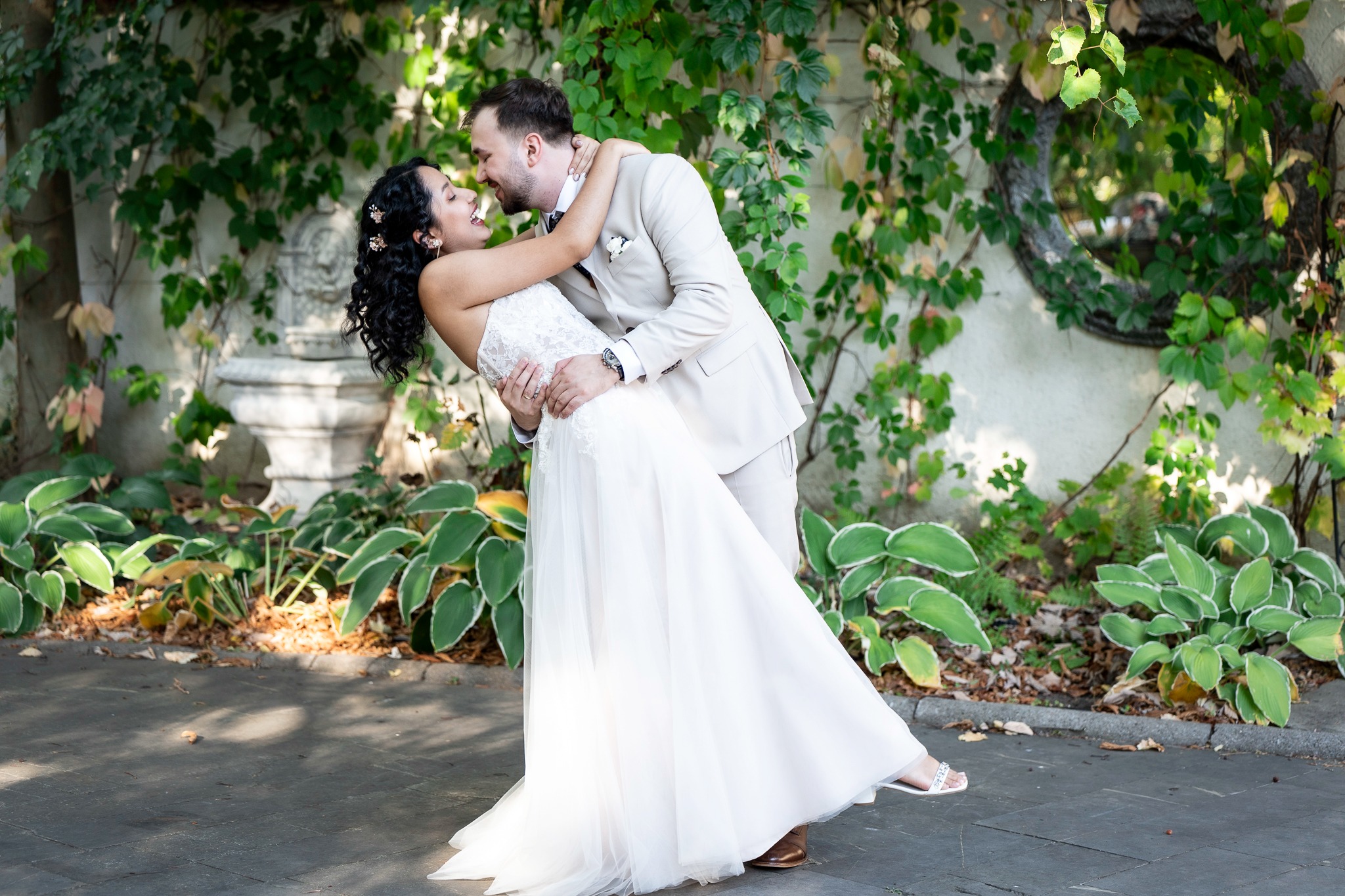 Uśmiechnięta para młoda w objęciach na tle zielonej ściany z bluszczu i dekoracyjnej fontanny. Panna młoda w białej sukni, pan młody w beżowym garniturze.