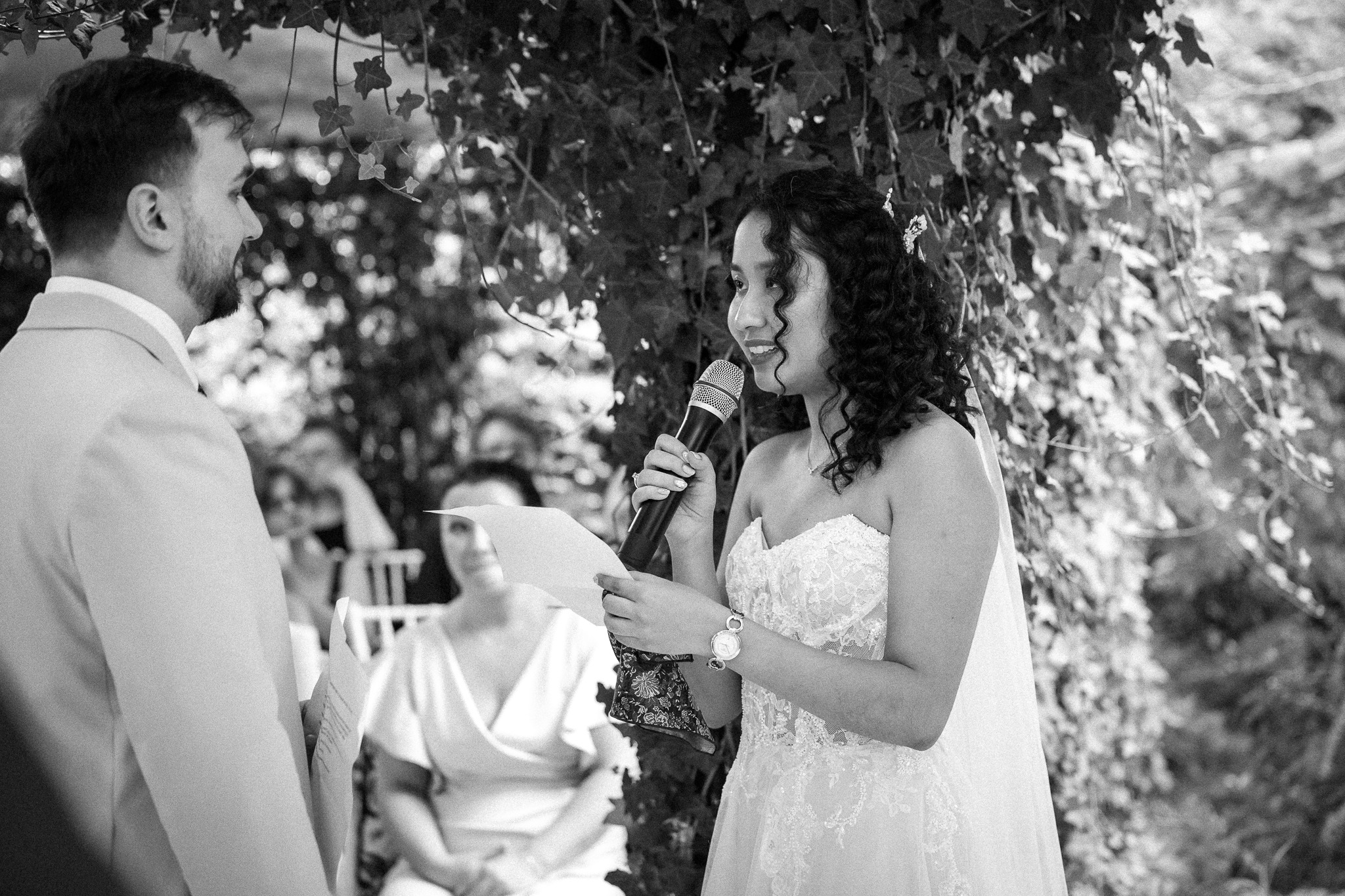 Czarno-białe zdjęcie panny młodej w koronkowej sukni, czytającej przysięgę do mikrofonu podczas ceremonii ślubnej na tle zieleni i gości.