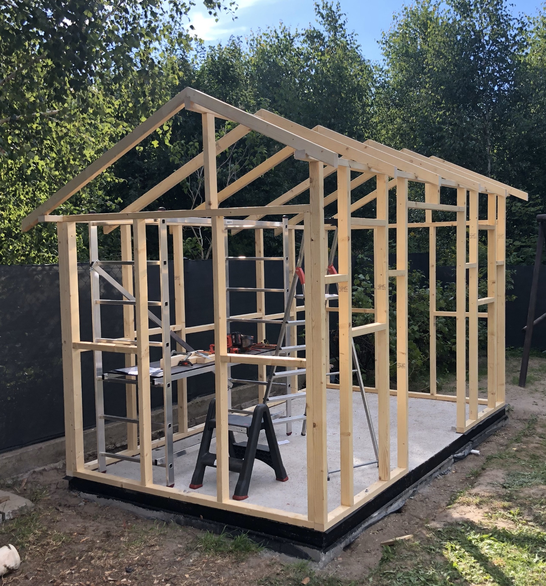Konstrukcja szkieletowa altany w trakcie budowy, widoczne drewniane elementy konstrukcyjne, betonowa podstawa, drabiny, narzędzia i zielone otoczenie.