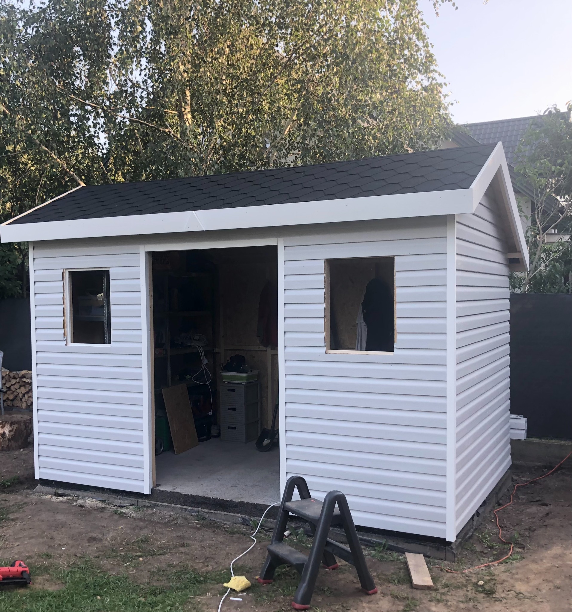 W trakcie budowy: Szopa ogrodowa z białym sidingiem, czarnym dachem z papy i ramami okiennymi bez szyb, widoczne wnętrze z narzędziami i drabina na zewnątrz.