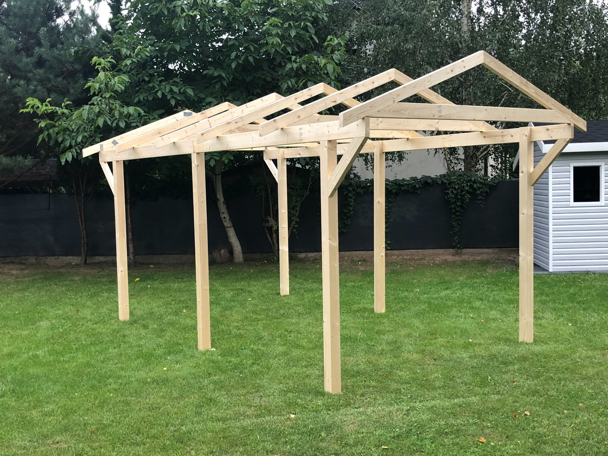 Konstrukcja z surowego drewna, prawdopodobnie w trakcie budowy, na zielonym trawniku, z widocznym małym budynkiem gospodarczym w tle.