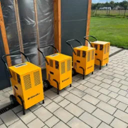 Cztery żółte osuszacze powietrza marki Riwali Pro ustawione rzędem na kostce brukowej przed budynkiem z szarą roletą i ścianą z folią budowlaną.