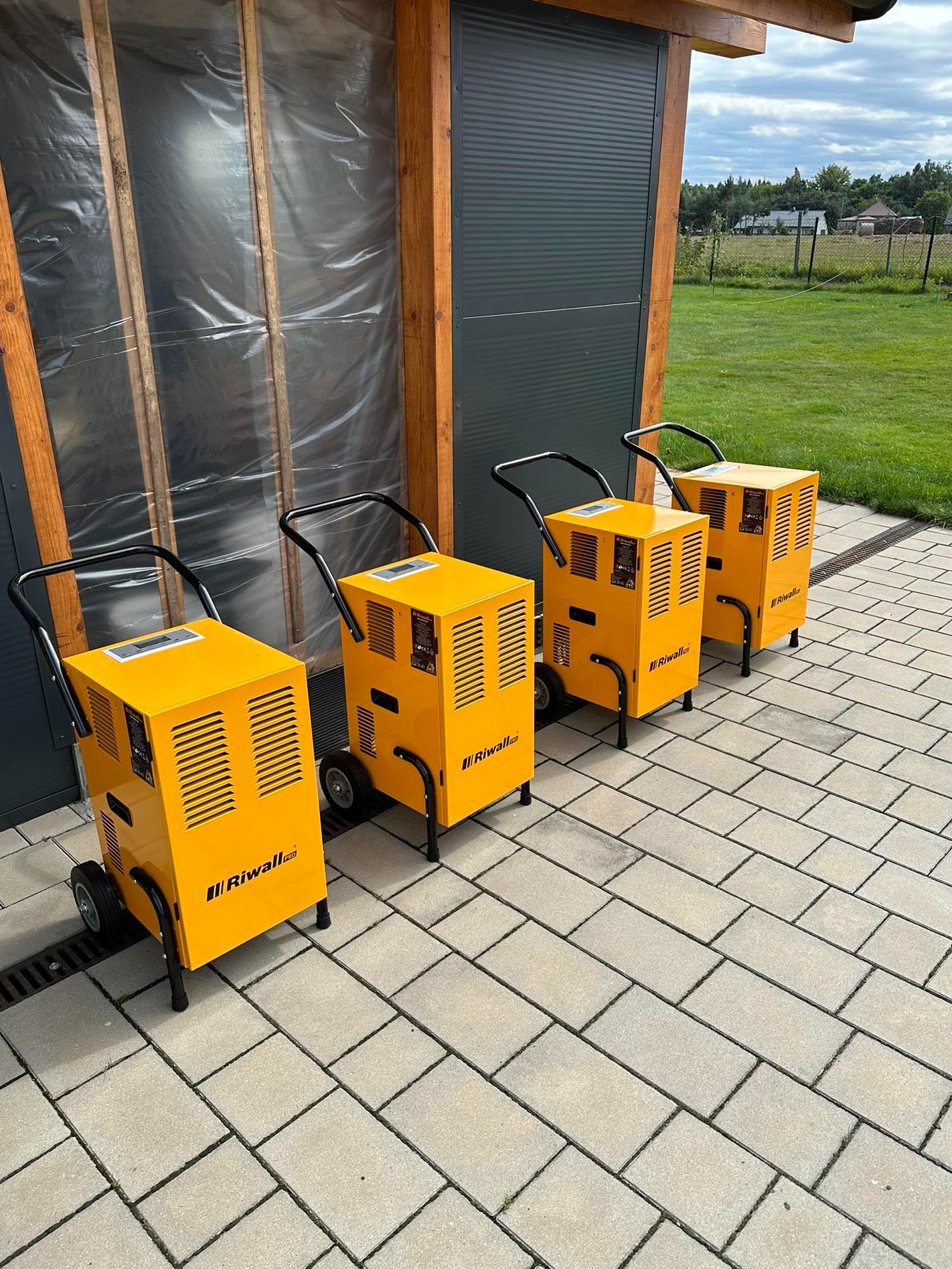 Cztery żółte osuszacze powietrza marki Riwali Pro ustawione rzędem na kostce brukowej przed budynkiem z szarą roletą i ścianą z folią budowlaną.