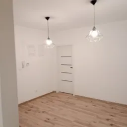 Wnętrze pokoju po remoncie: jasne panele podłogowe, białe ściany, białe drzwi z czarnymi akcentami i dwie geometryczne lampy wiszące.