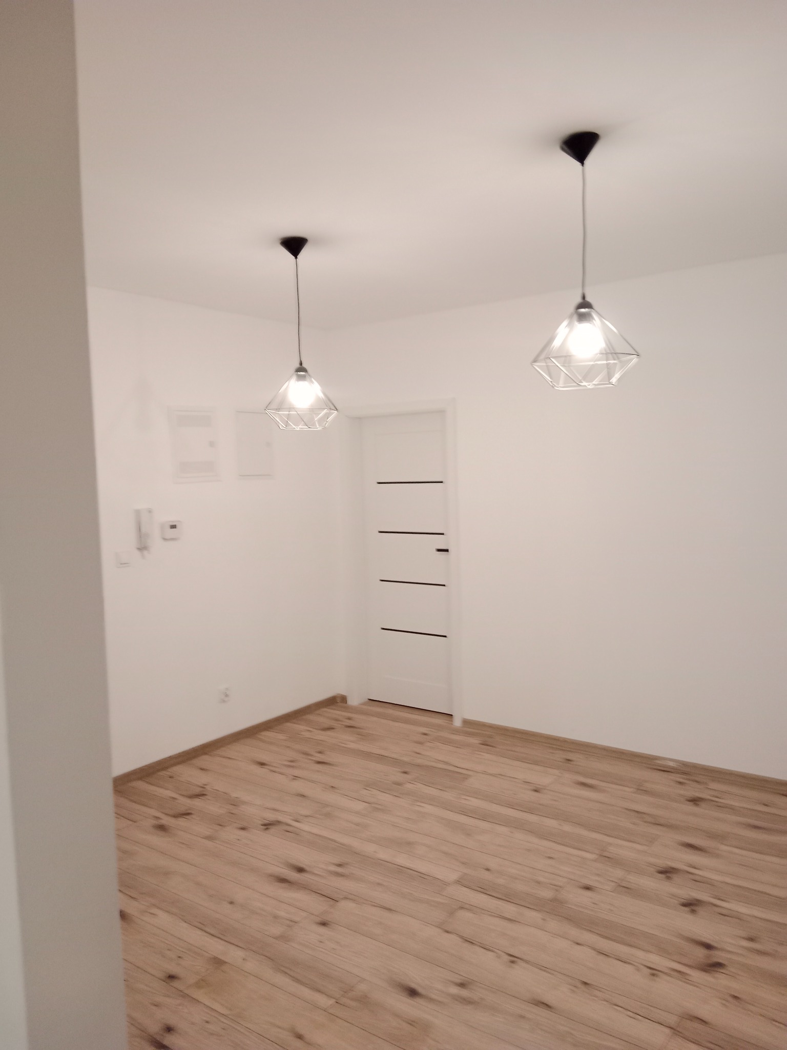 Wnętrze pokoju po remoncie: jasne panele podłogowe, białe ściany, białe drzwi z czarnymi akcentami i dwie geometryczne lampy wiszące.