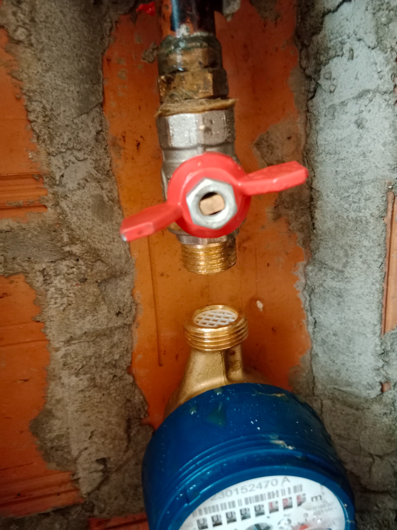 Fragment instalacji wodnej z zaworem z czerwoną rączką i licznikiem wody z niebieską obudową, zamontowany na surowej ścianie z cegły i betonu.