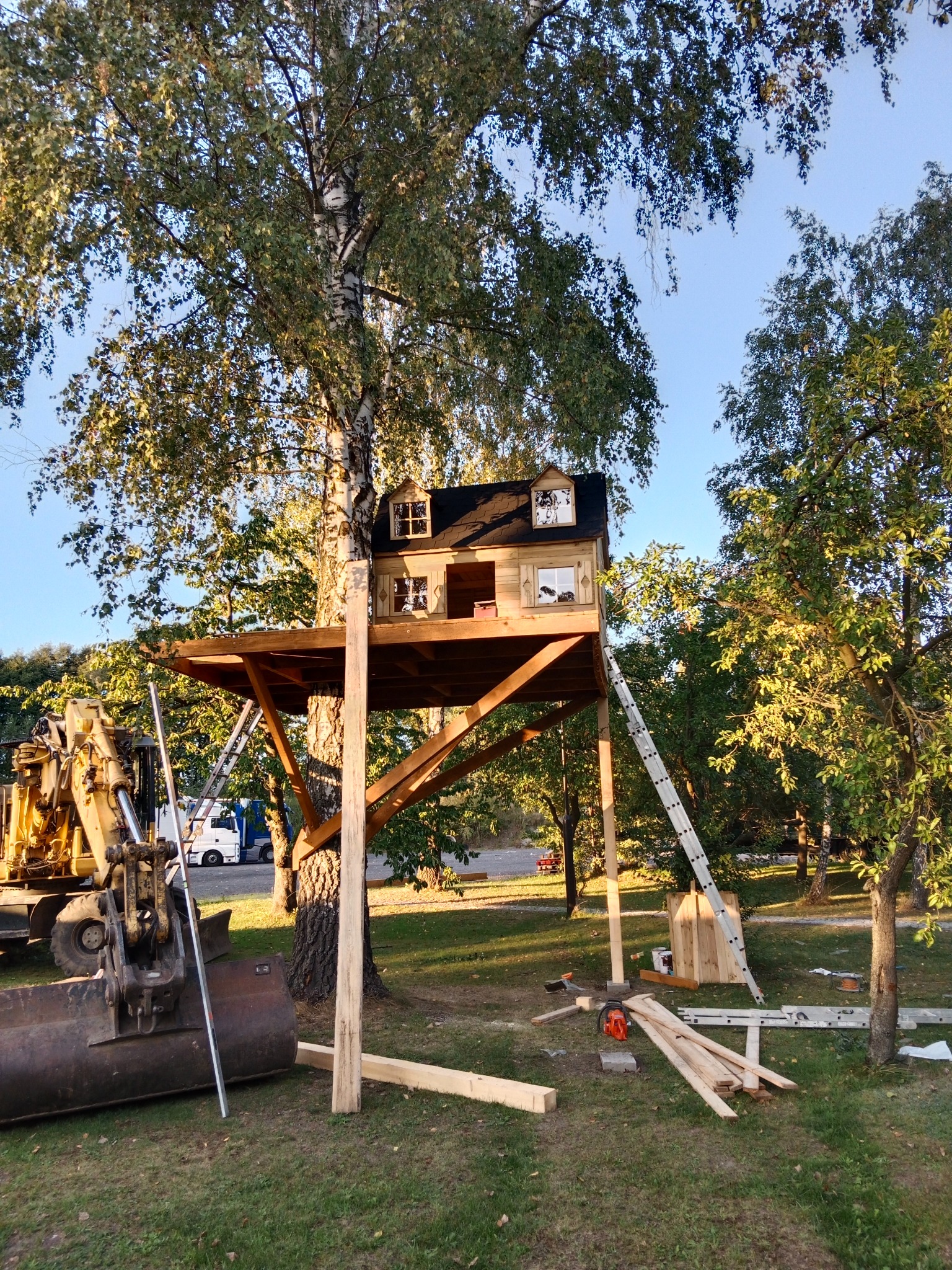Budowa domku na drzewie: drewniana konstrukcja na platformie, z elementami wykończeniowymi, drabina oparta o platformę, na trawie leżą narzędzia i deski, w tle koparka i samochody.