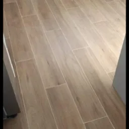 Ułożona podłoga z płytek imitujących drewno w jasnobrązowym odcieniu, widoczne białe fugi, w tle białe szafki kuchenne.