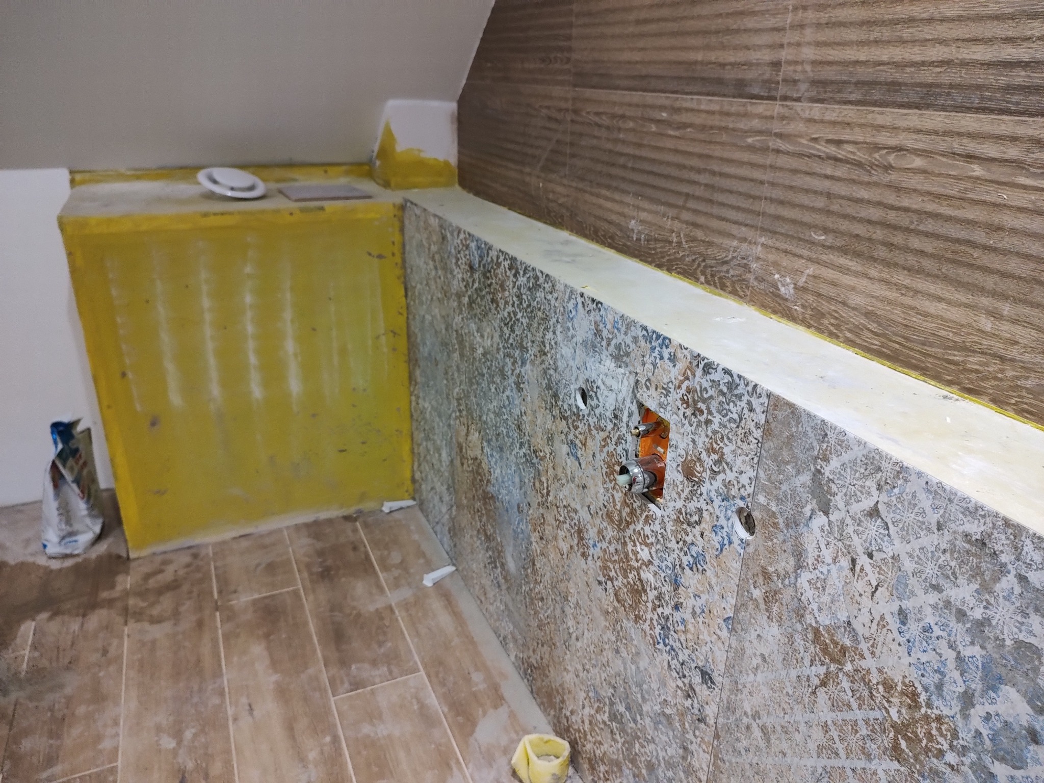 Remont łazienki w trakcie: montaż płytek z wzorem na ścianie, widoczne przyłącza wodne, żółta zabudowa pod umywalkę, podłoga wyłożona płytkami imitującymi drewno.