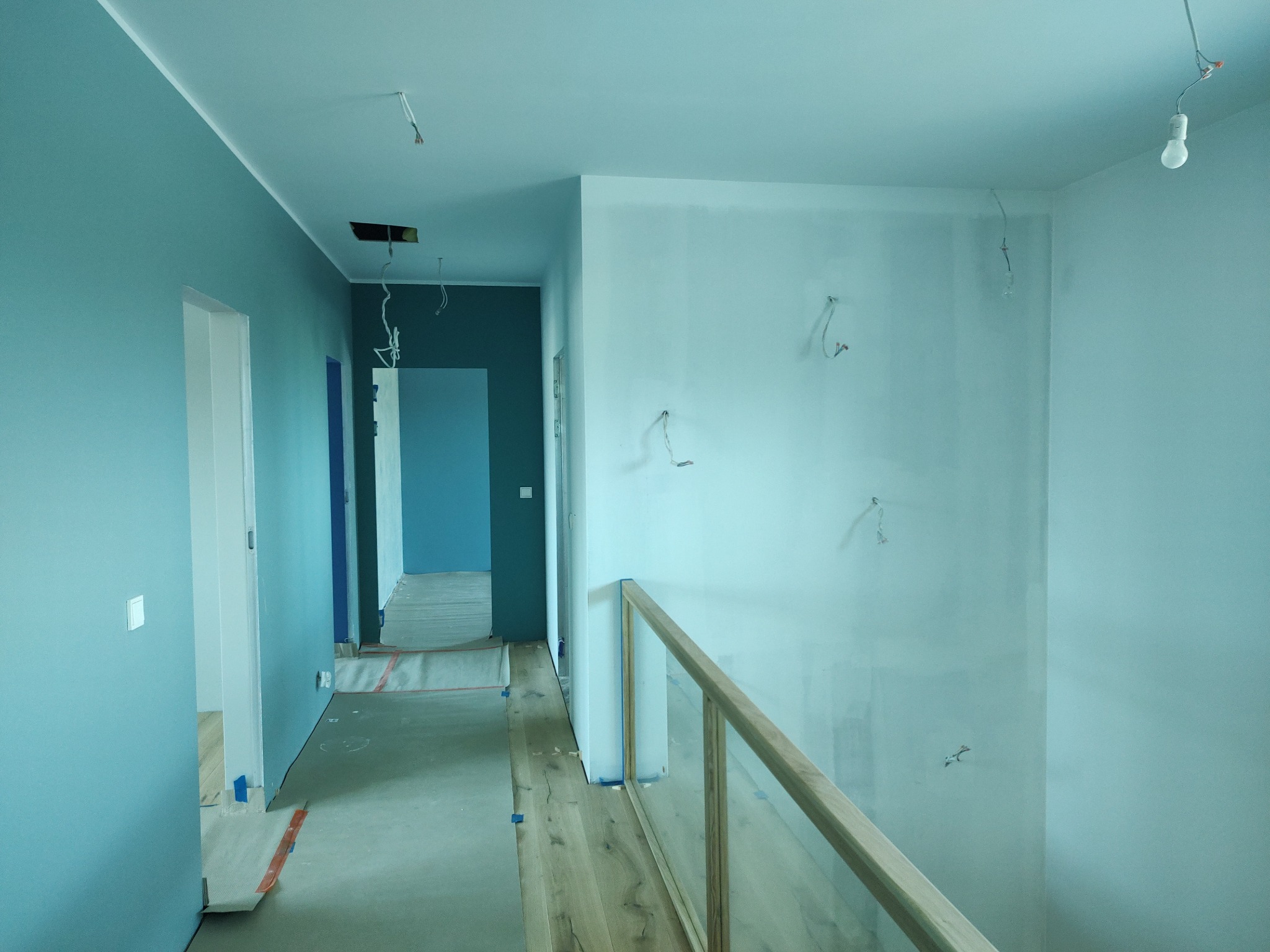Korytarz w trakcie remontu z pomalowanymi na niebiesko ścianami, widoczne przewody elektryczne wystające ze ścian i sufitu, podłoga zabezpieczona folią malarską, drewniana balustrada z elementami...