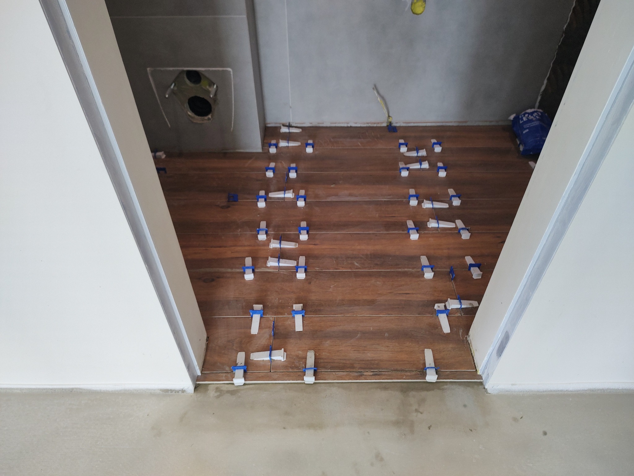 Układanie brązowych płytek podłogowych imitujących drewno w łazience, z użyciem białych i niebieskich krzyżyków dystansowych, widoczne szare ściany i otwór wentylacyjny.