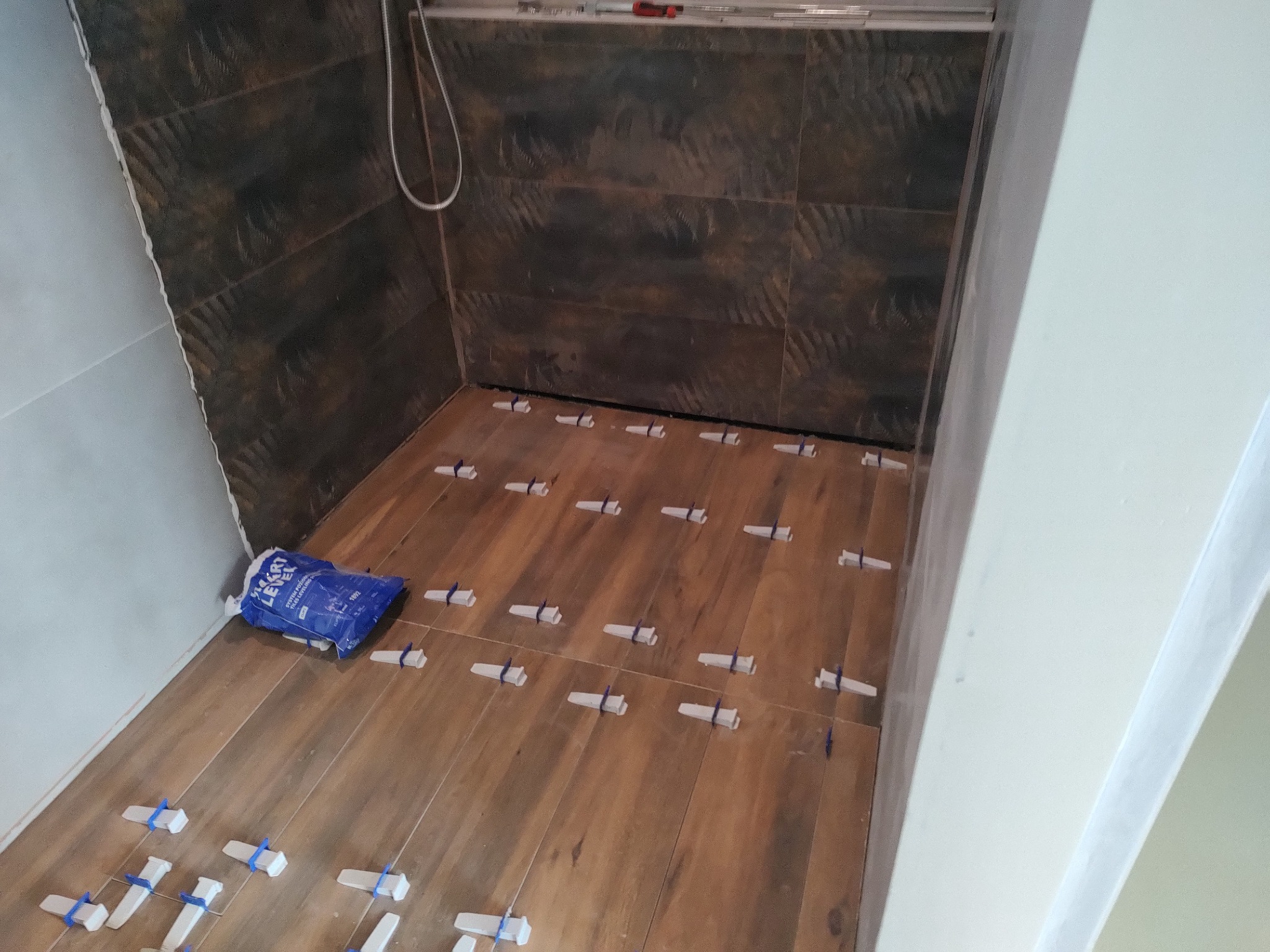 Układanie brązowych płytek podłogowych imitujących drewno w łazience, z użyciem systemu poziomowania płytek z niebieskimi klinami, obok leży worek z klejem do płytek.