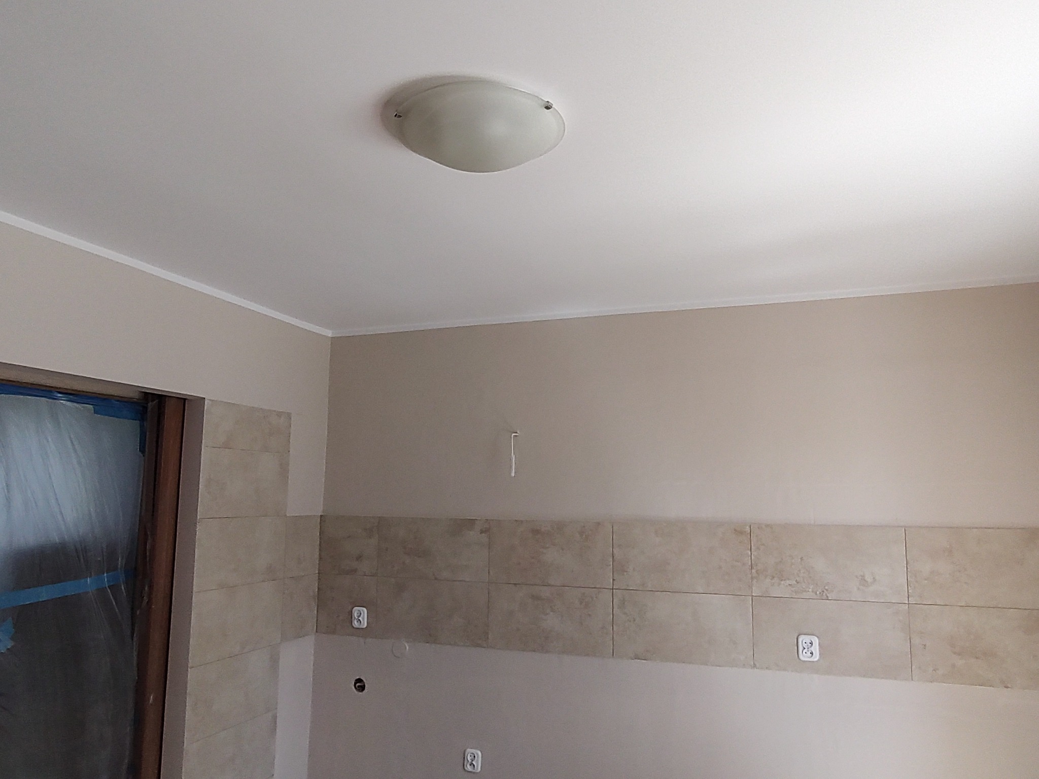 Ściana łazienki z beżowymi płytkami, widoczne gniazdka elektryczne i zabezpieczone folią drzwi, sufit z lampą.