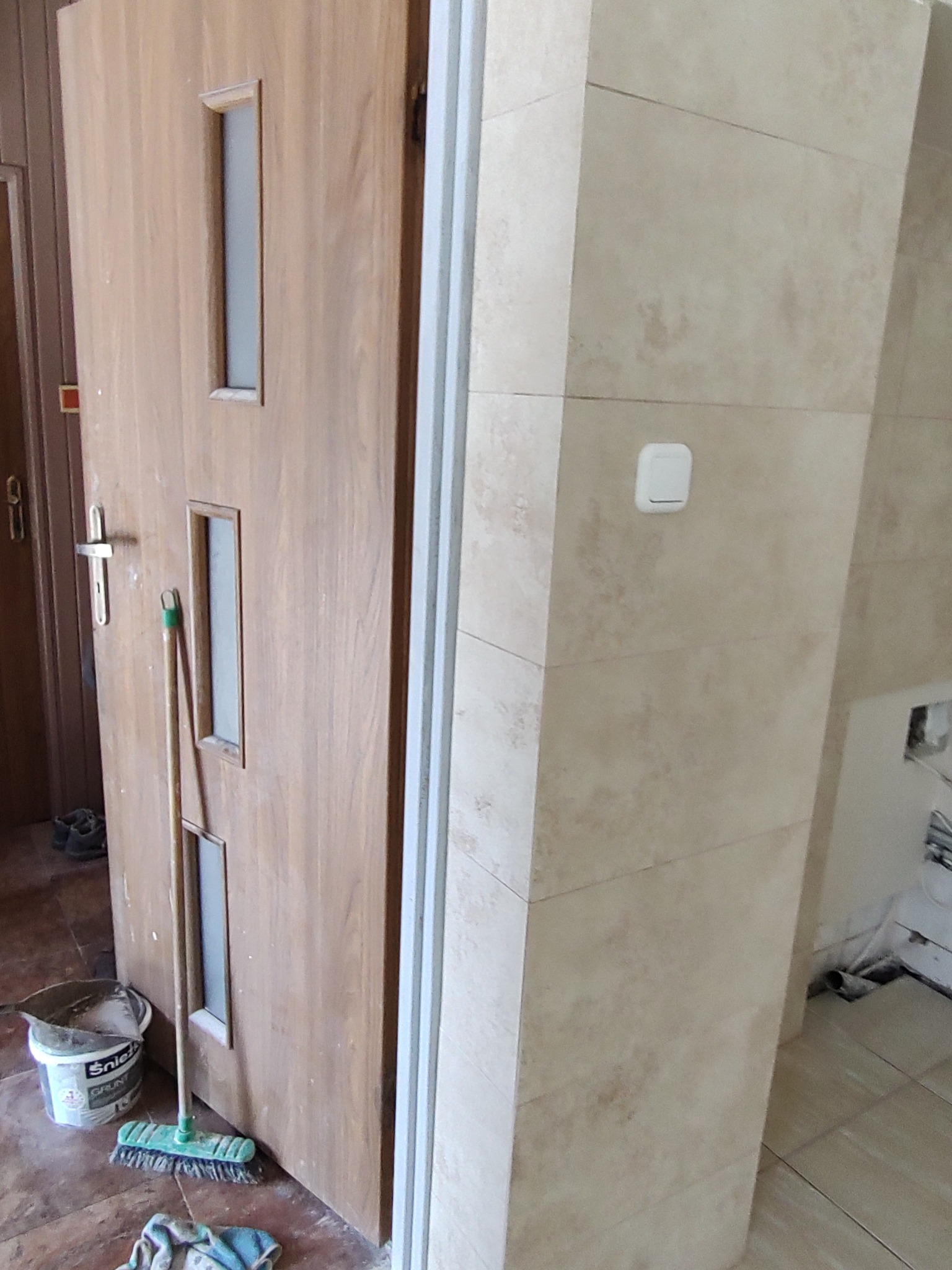 Drewniane drzwi z trzema szybkami w trakcie montażu, widoczna ościeżnica i fragment wykończonej ściany z beżowych płytek ceramicznych, na podłodze wiadro z gruntem i szczotka