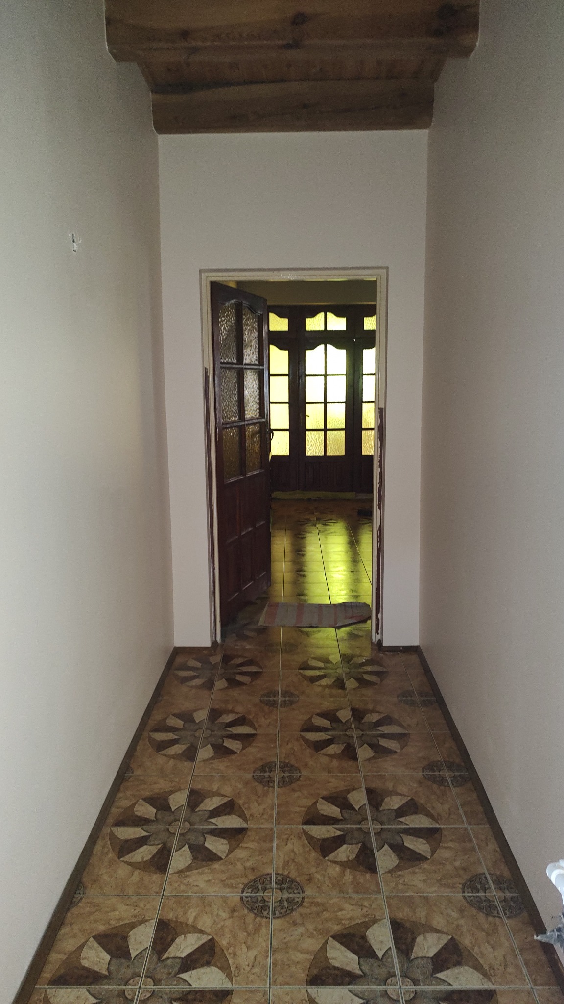 Wykończony korytarz z brązowymi płytkami podłogowymi w kwiatowe wzory, drewnianą belką stropową i otwartymi, przeszklonymi drzwiami do kolejnego pomieszczenia z żółtym światłem.