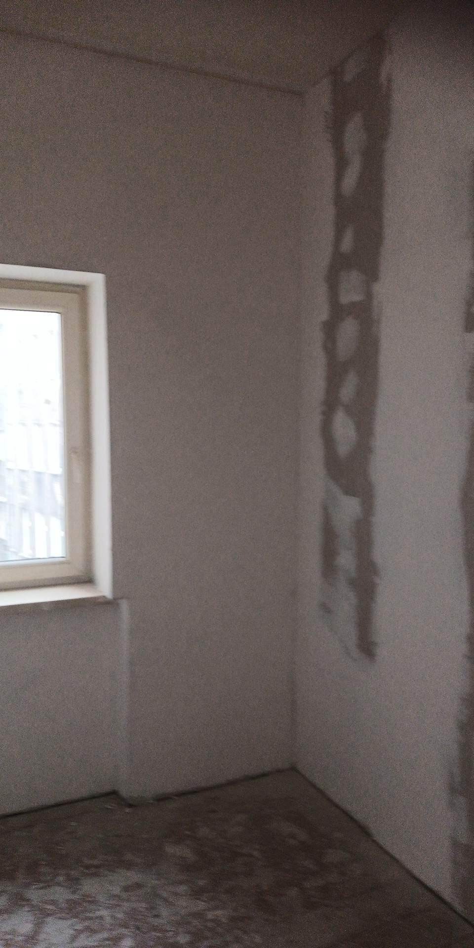 Surowe ściany w pokoju podczas remontu, widoczne ślady po położeniu gładzi gipsowej, okno po lewej stronie kadru, przygotowanie do dalszych prac wykończeniowych.