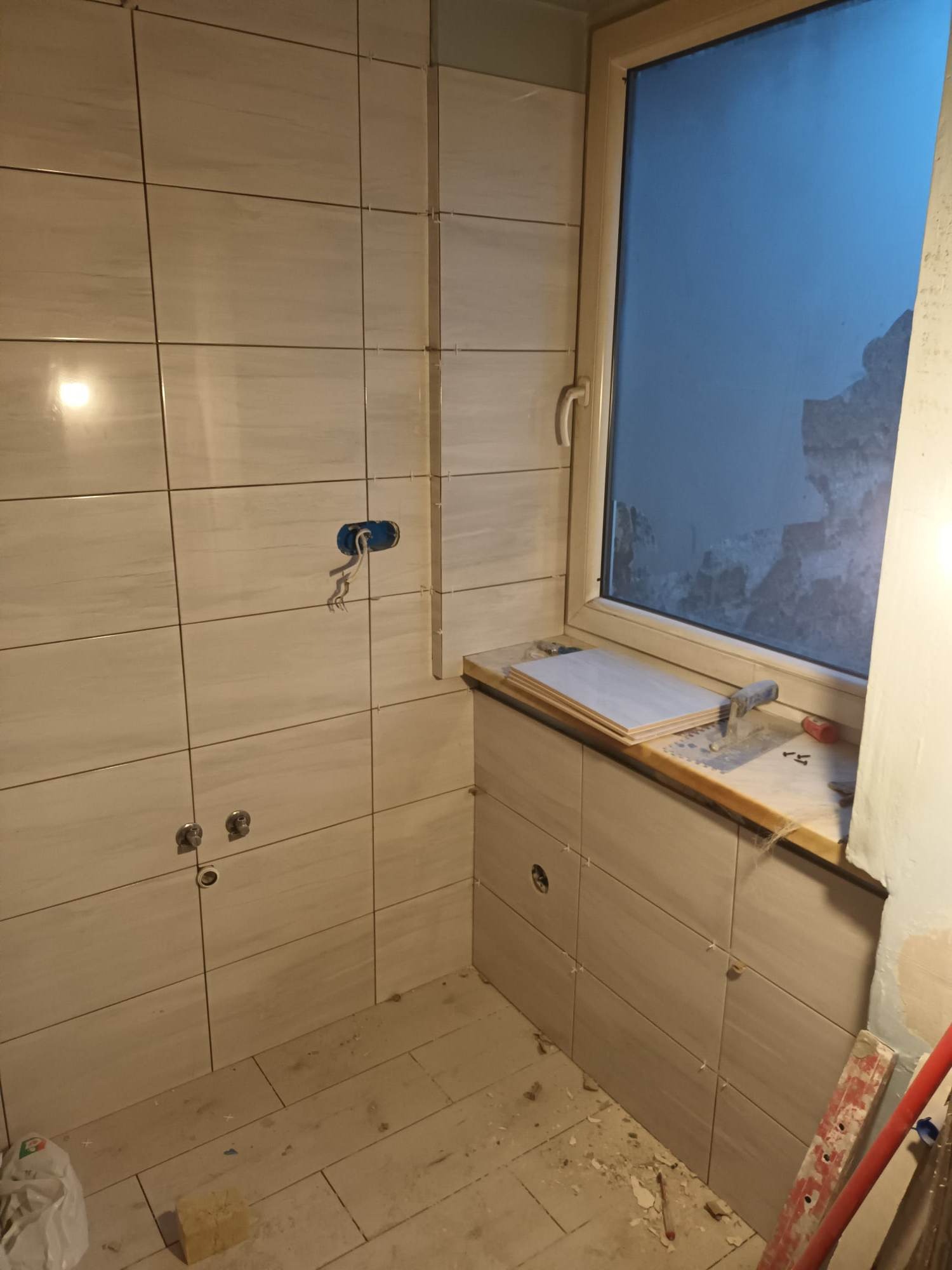 Ściana łazienki w trakcie układania płytek ceramicznych, widoczne krzyżyki dystansowe, puszka elektryczna z wystającymi przewodami oraz okno z widokiem na zewnątrz. Na parapecie leżą płytki...