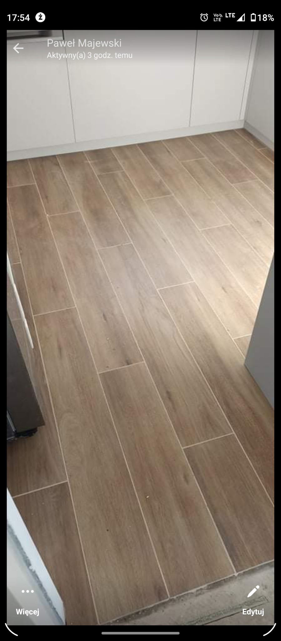 Ułożona podłoga z płytek imitujących drewno w jasnobrązowym odcieniu, widoczne białe fugi, w tle białe szafki kuchenne.