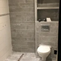 Łazienka w trakcie wykańczania: szare płytki imitujące kamień na ścianach i podłodze, zabudowana półka w ścianie, wisząca toaleta i odpływ liniowy w podłodze, widoczna skrzynka z narzędziami.