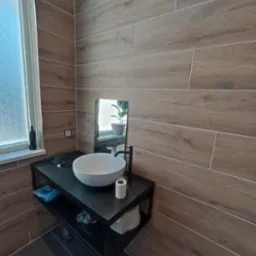 Nowoczesna łazienka z drewnopodobnymi płytkami na ścianach, okrągłą białą umywalką nablatową na czarnym metalowym regale i czarną armaturą.