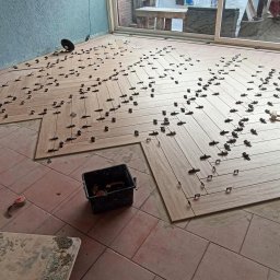 Tanio i z fakturką - Układanie podłogi z płytek imitujących drewno w jodełkę, z użyciem klipsów poziomujących, widoczny fragment starej podłogi i pojemnik z narzędziami.